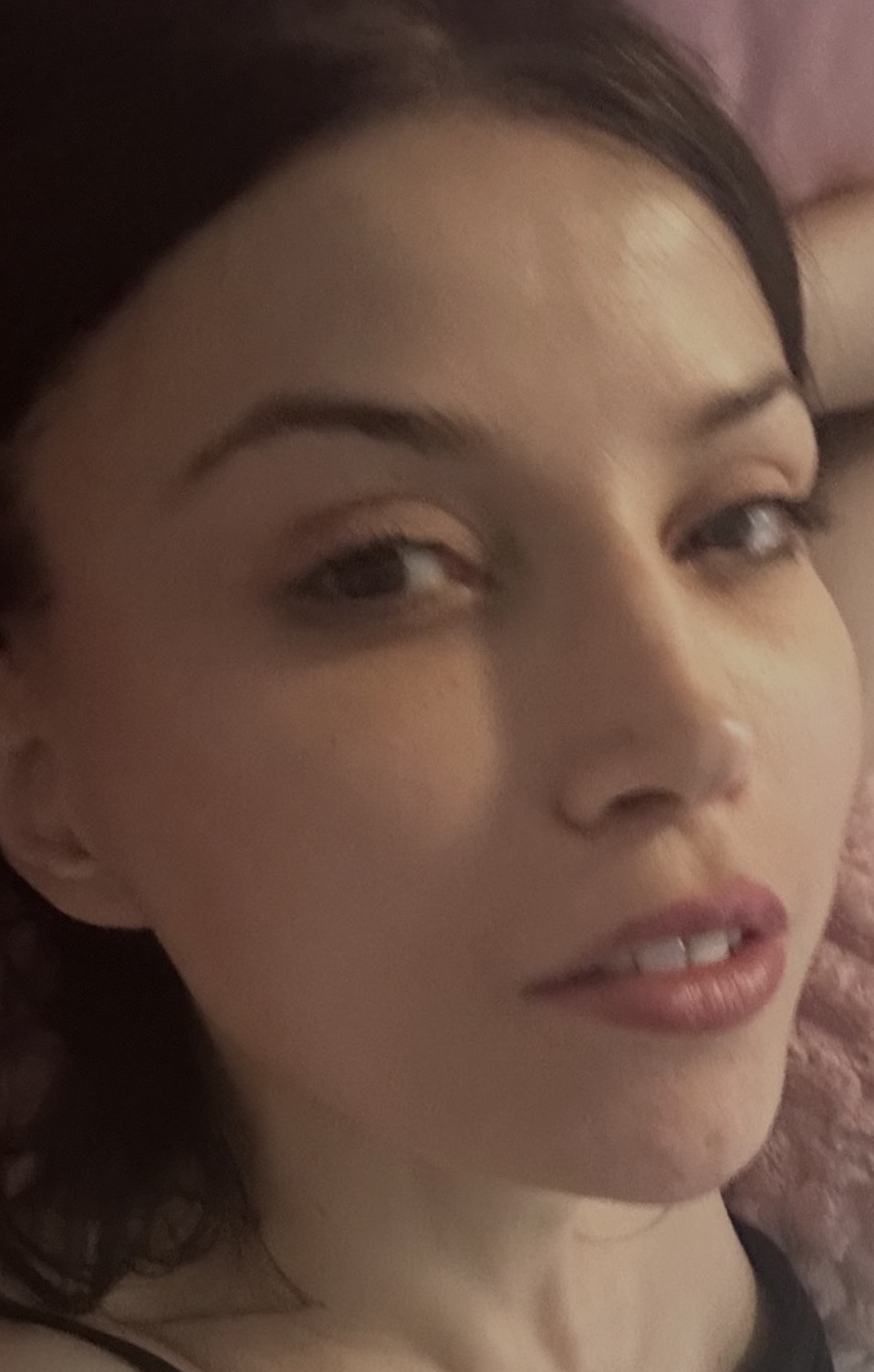 Close-up of a woman's face with dark hair, lying on a pink pillow, looking at the camera.