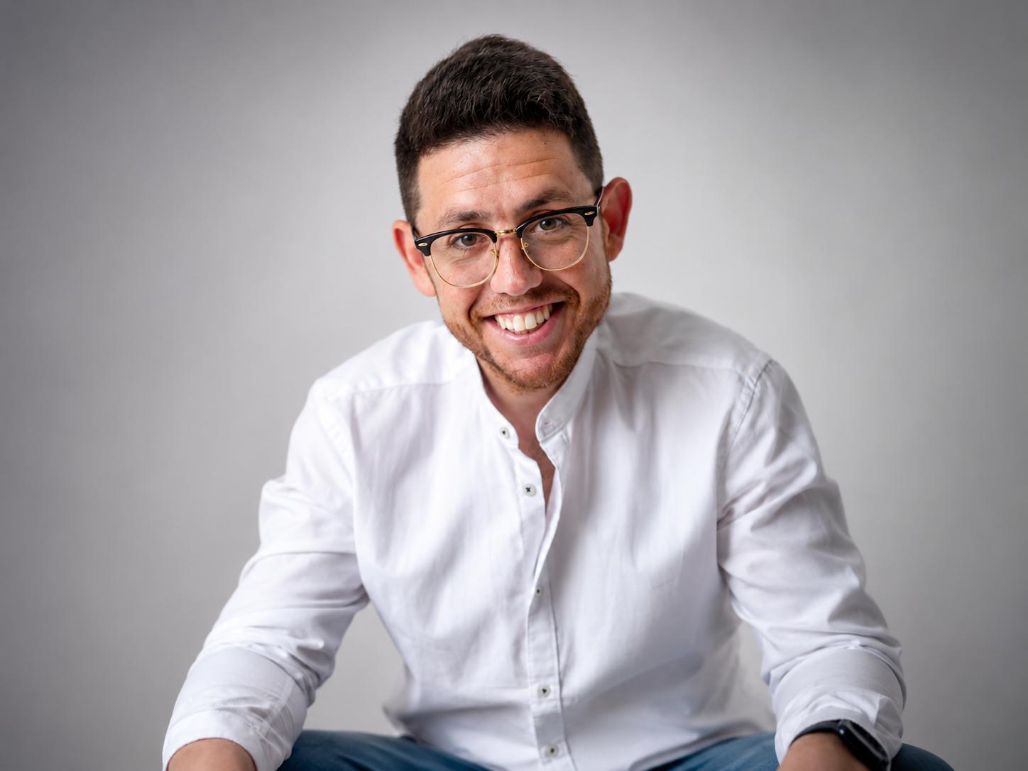 Smiling man with glasses in a white shirt against a plain background.