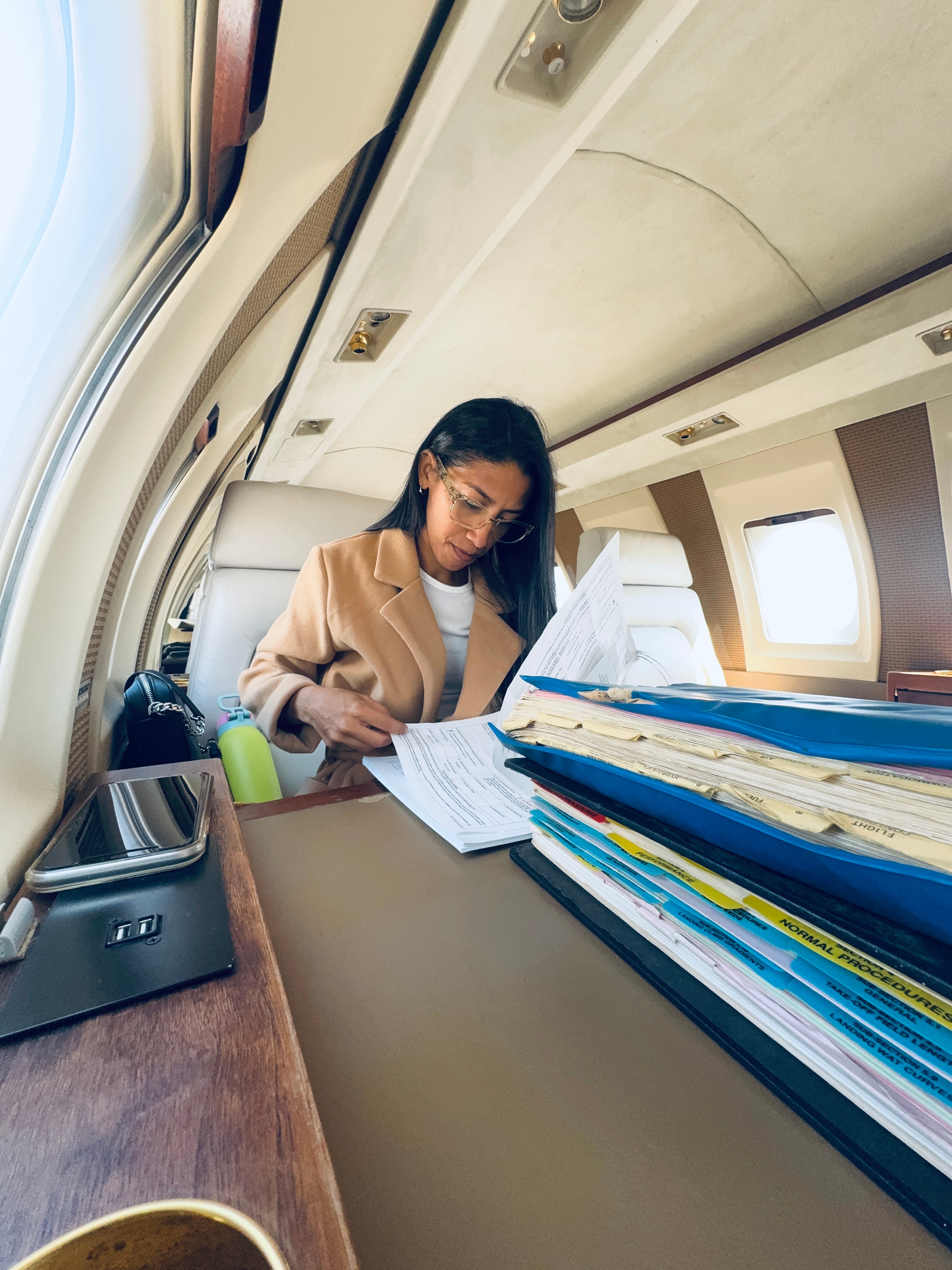 Woman sitting at a desk inside an airplane, working with papers and files, window and overhead compartments visible.