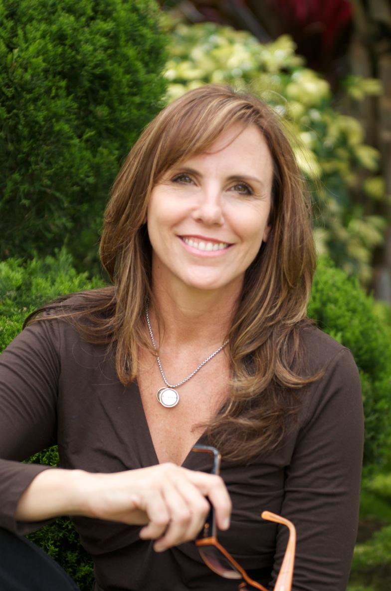 Smiling woman with brown hair outdoors, surrounded by greenery, wearing a dark top and a necklace.