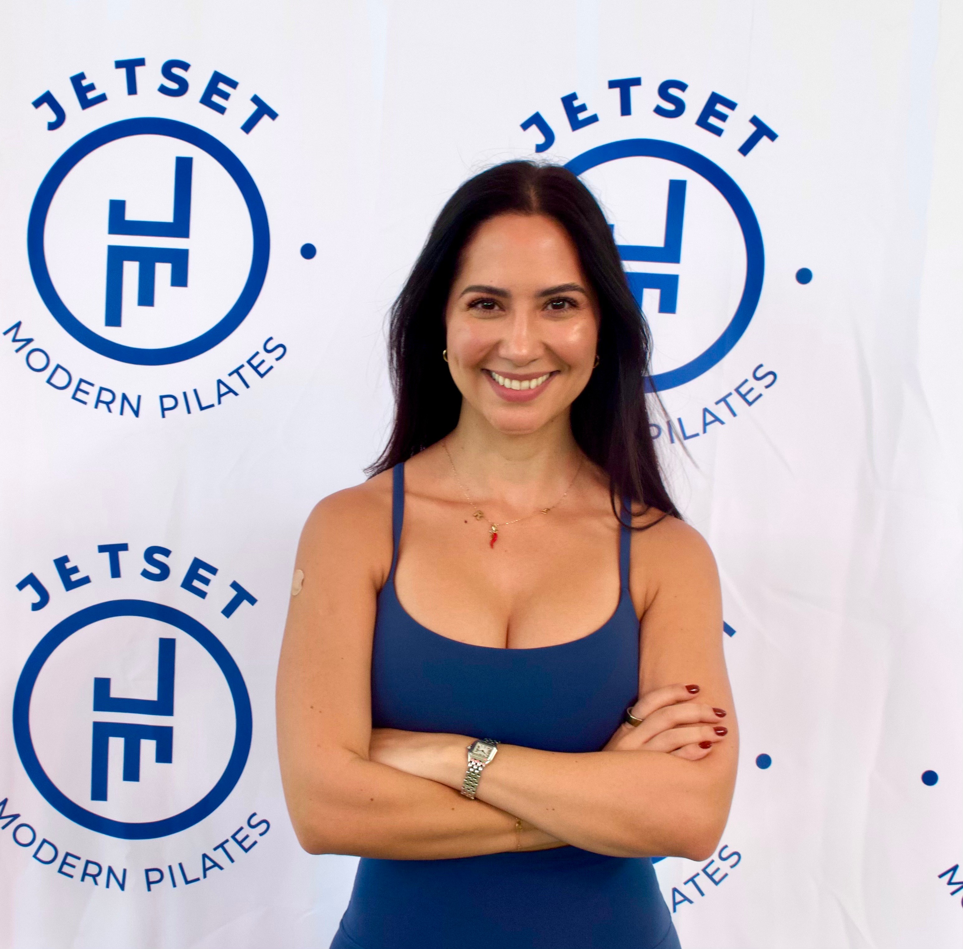Woman with dark hair smiling, arms crossed, standing in front of Jetset Modern Pilates logo backdrop.