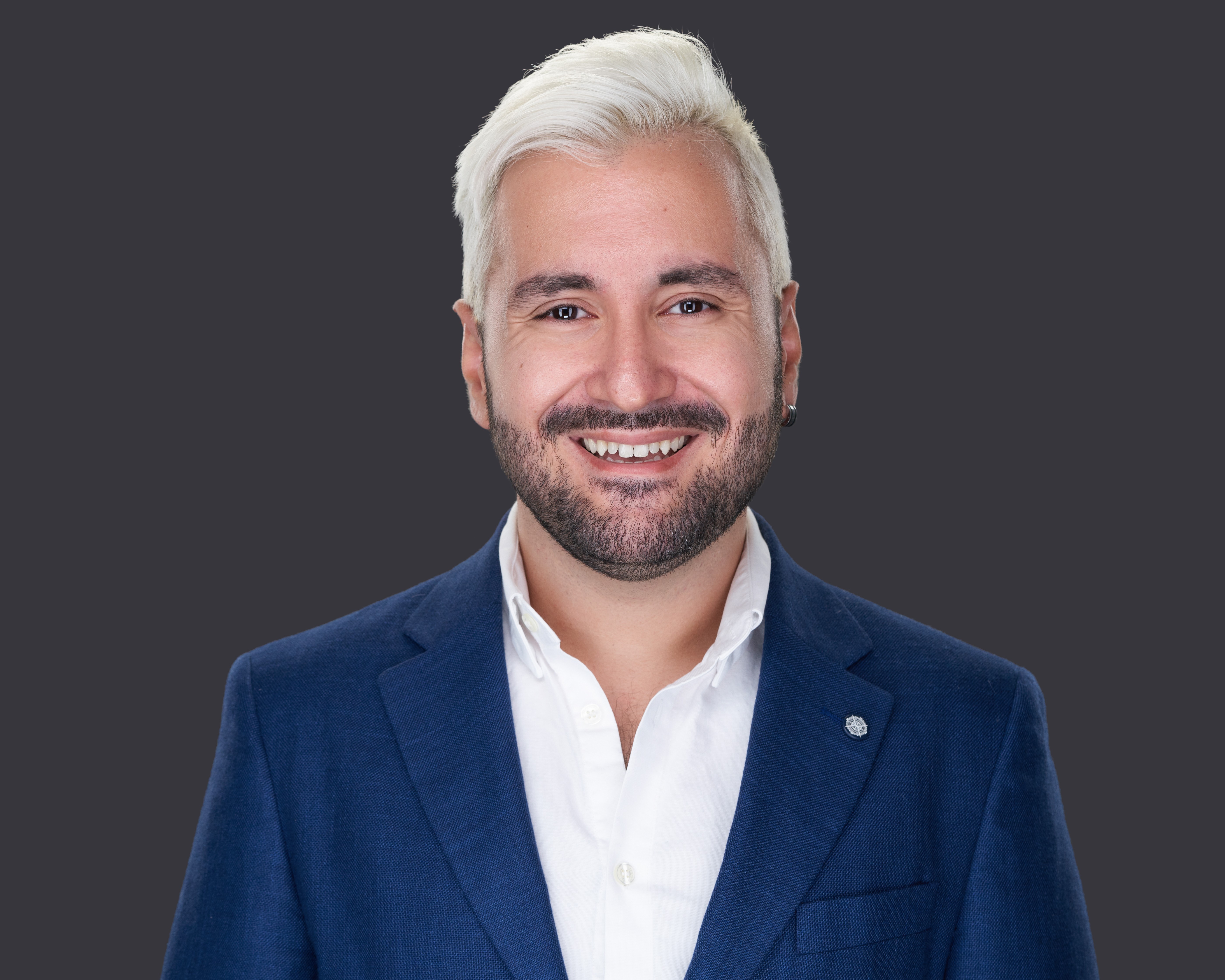 Portrait of a smiling man with white hair and beard wearing a blue suit and white shirt against a dark background.