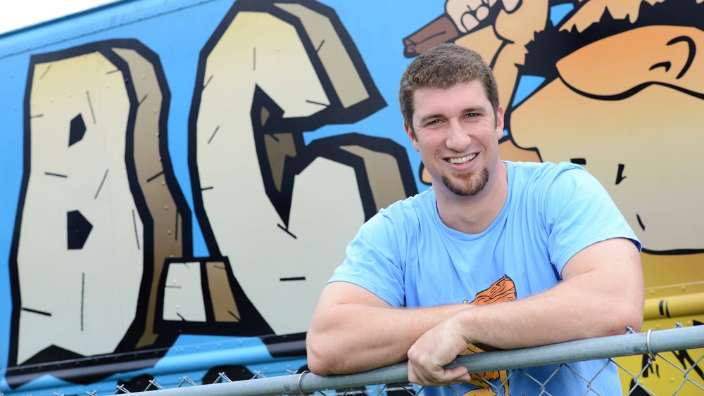 Young man with short hair and a beard smiling, wearing a light blue shirt, leaning on a yellow railing in front of graffiti art.