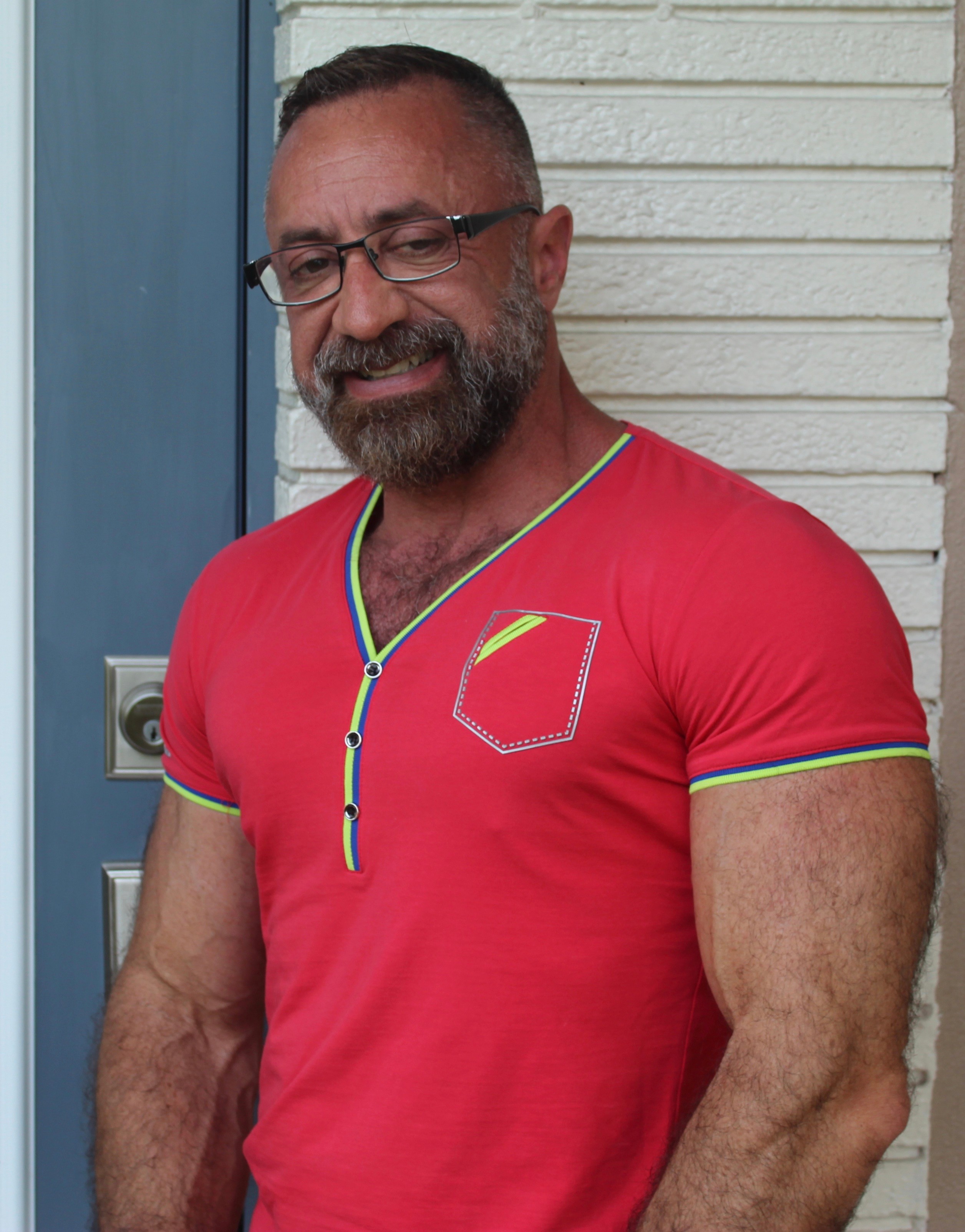 Man with glasses and beard wearing a red shirt with green and yellow trim, standing outdoors near a white wall.