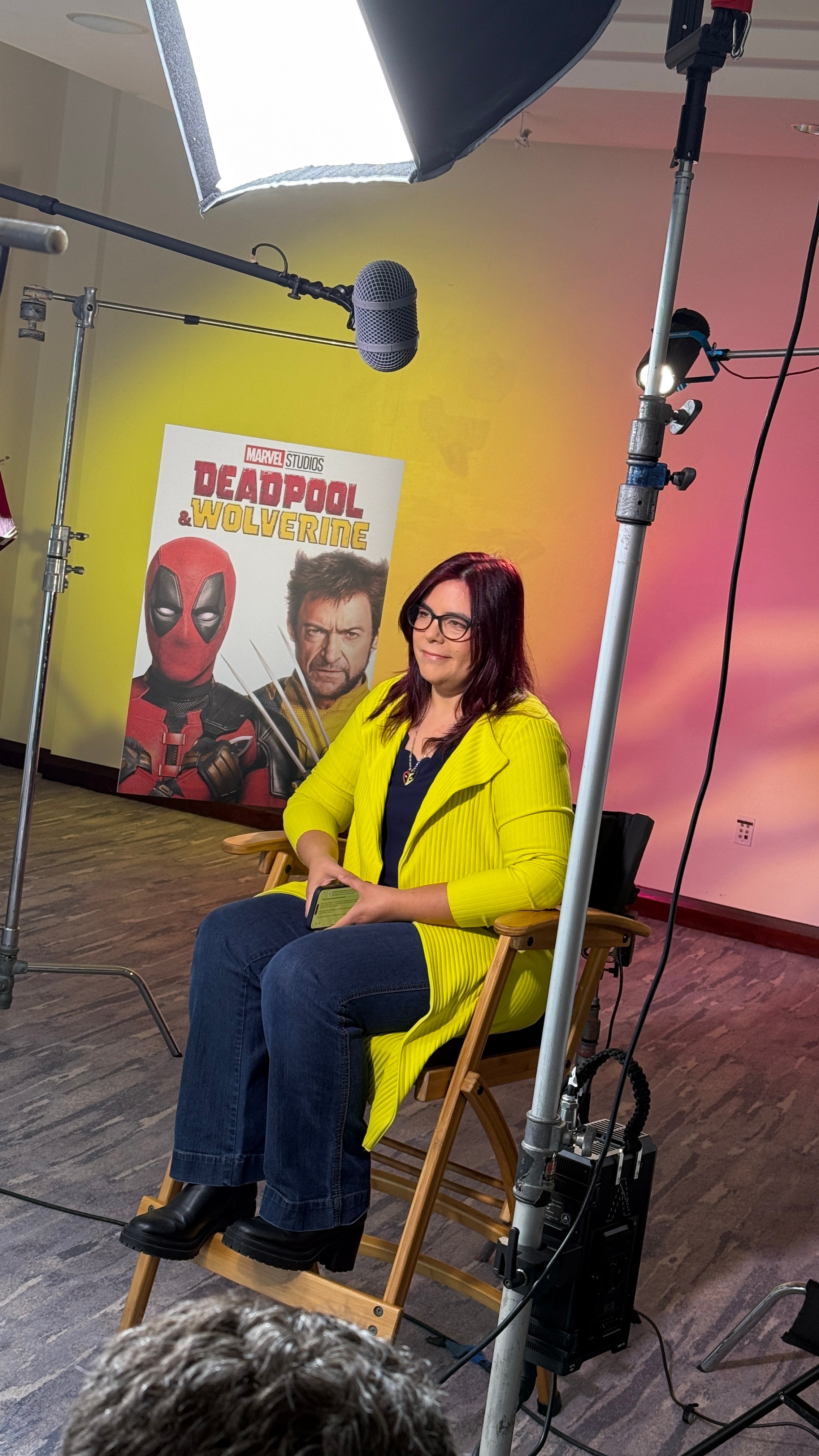 Woman with glasses and dark hair, wearing a yellow jacket, sitting on a chair in front of a Deadpool poster, studio lighting setup visible.