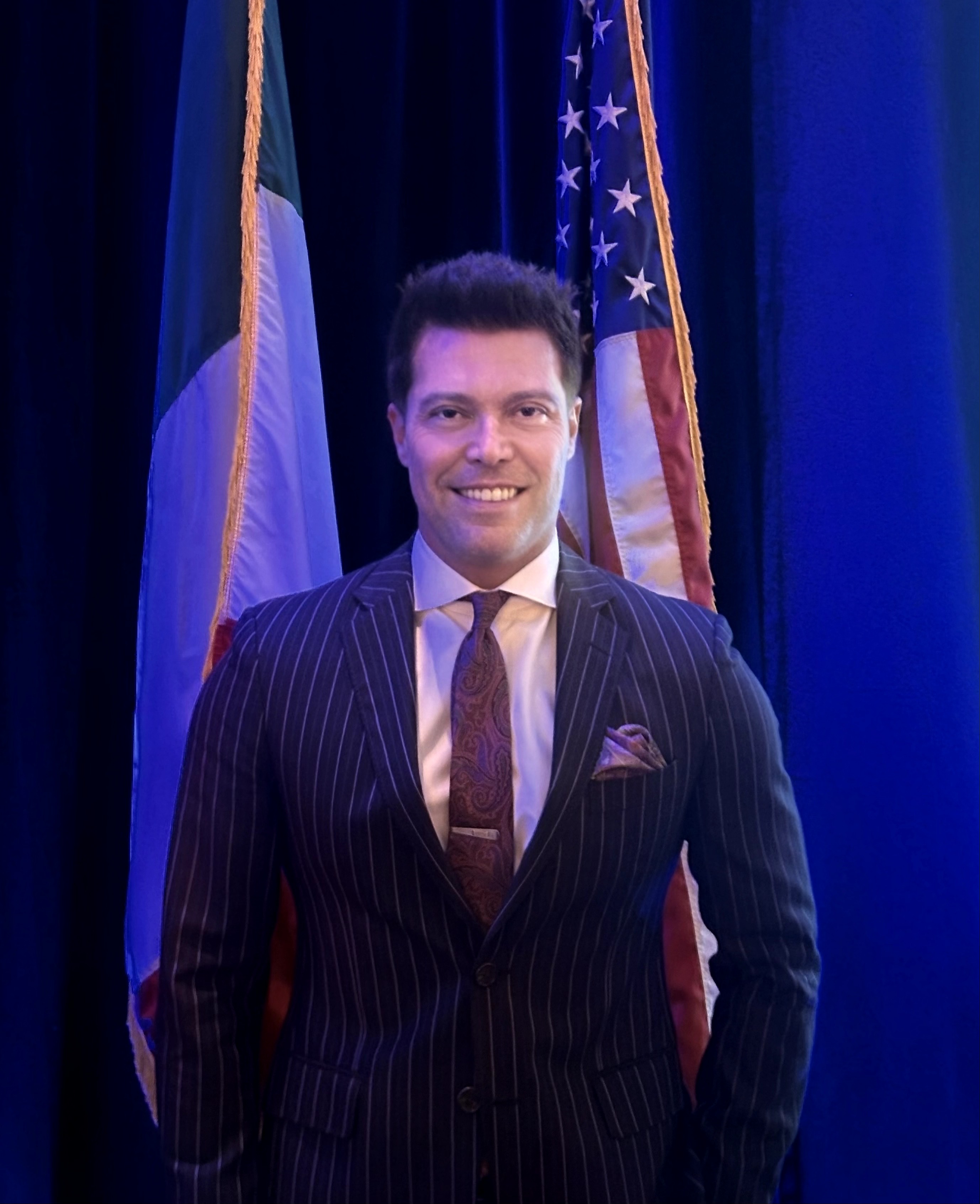 Man in a pinstripe suit standing in front of flags, smiling, with blue background.