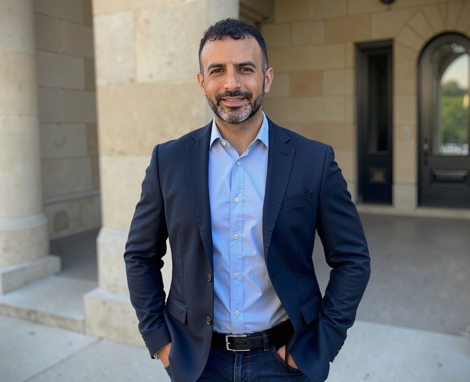 Man in a suit standing outdoors in front of a stone building, smiling, hands in pockets.