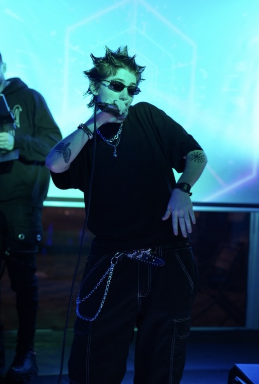 Person with spiky hair, sunglasses, and jewelry poses in a neon-lit environment, wearing a black outfit with chains.