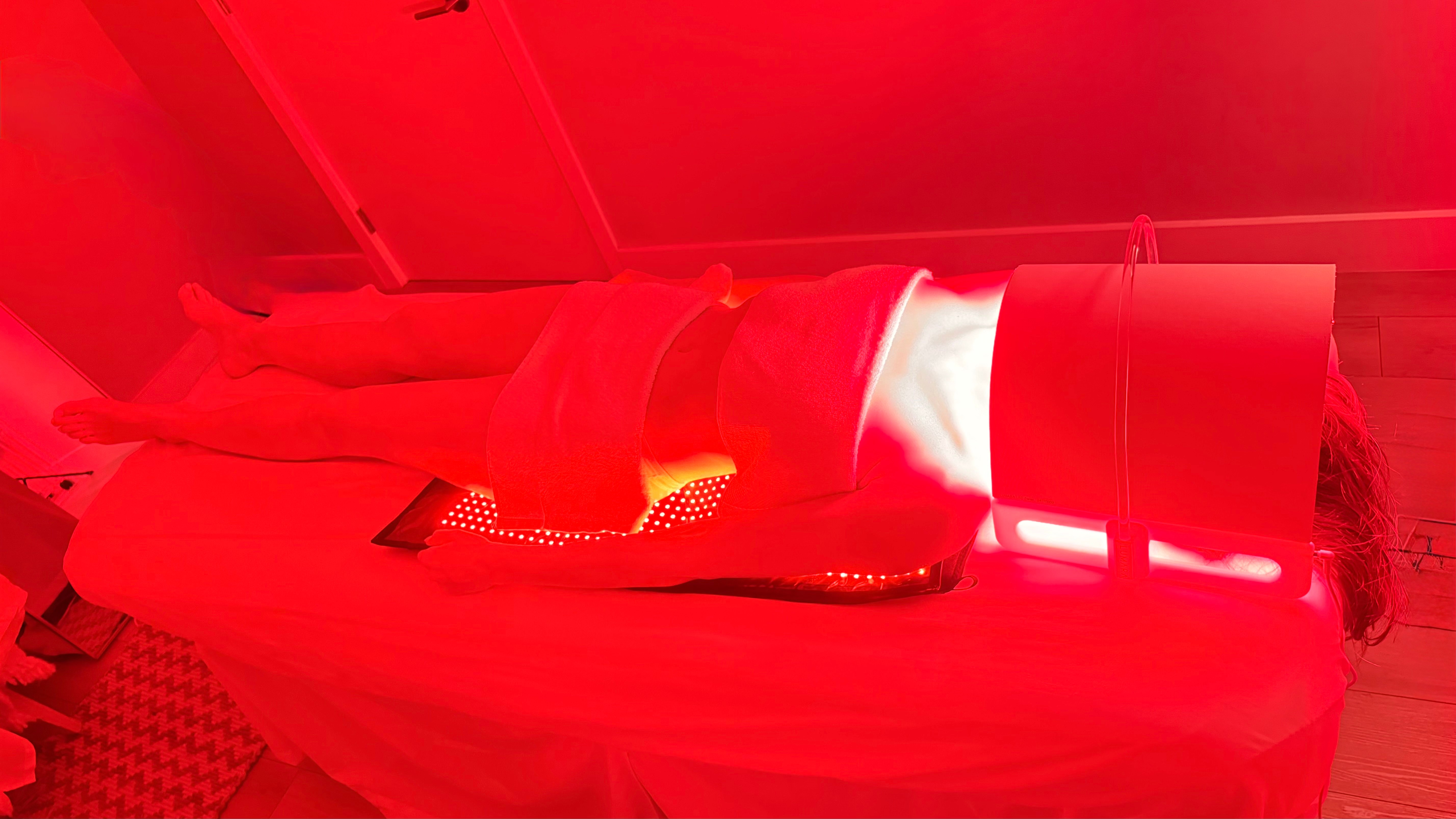 Person lying on a bed with a blanket, illuminated by red light, in a room with a ceiling and wall visible.