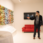 Person with long hair, black hat, and dark clothing points at a wall-mounted TV in a white gallery room with colorful art installation.