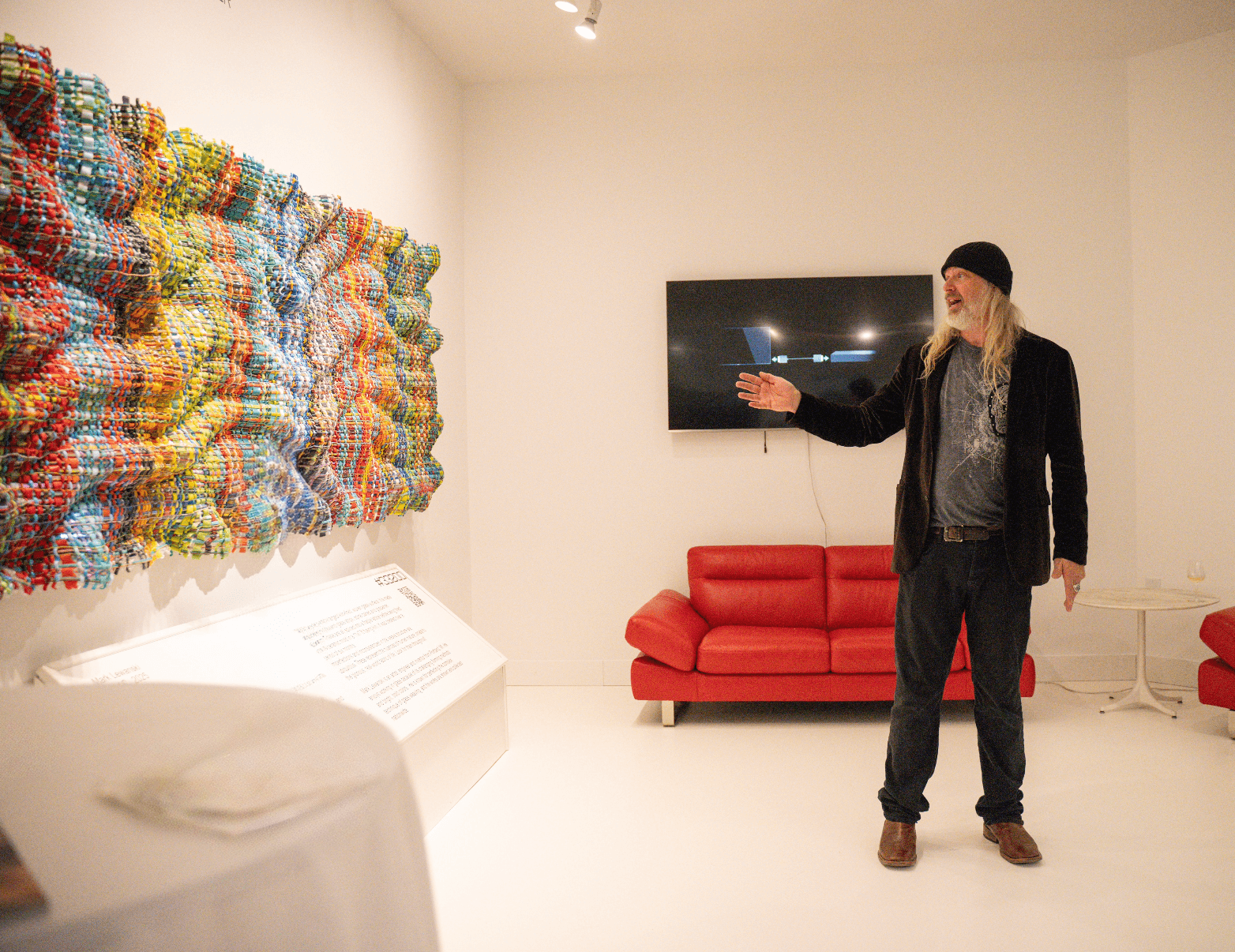 Person with long hair, black hat, and dark clothing points at a wall-mounted TV in a white gallery room with colorful art installation.
