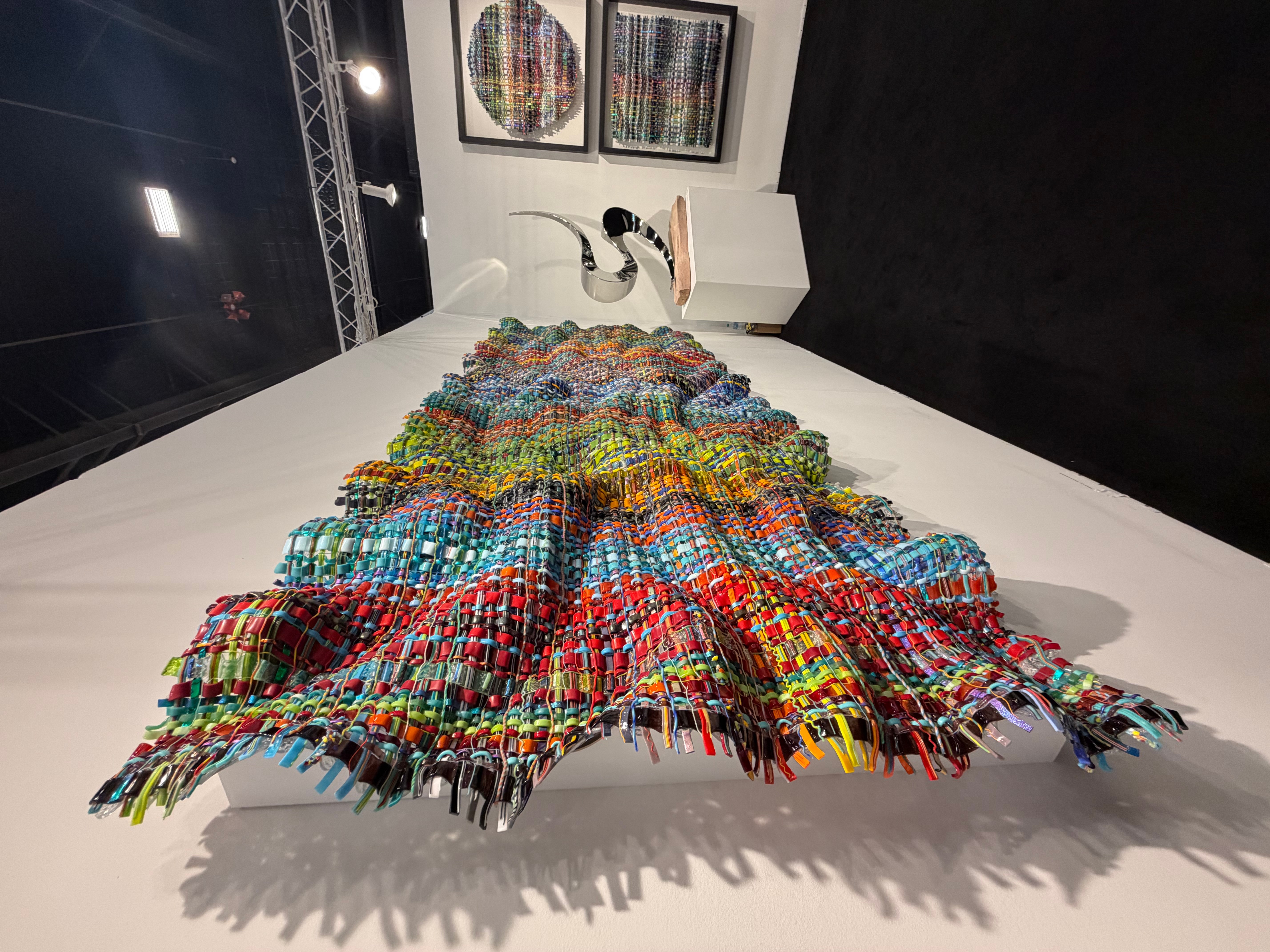 Colorful woven textile draped over a white table, with framed artwork on the wall behind, in an art gallery setting.
