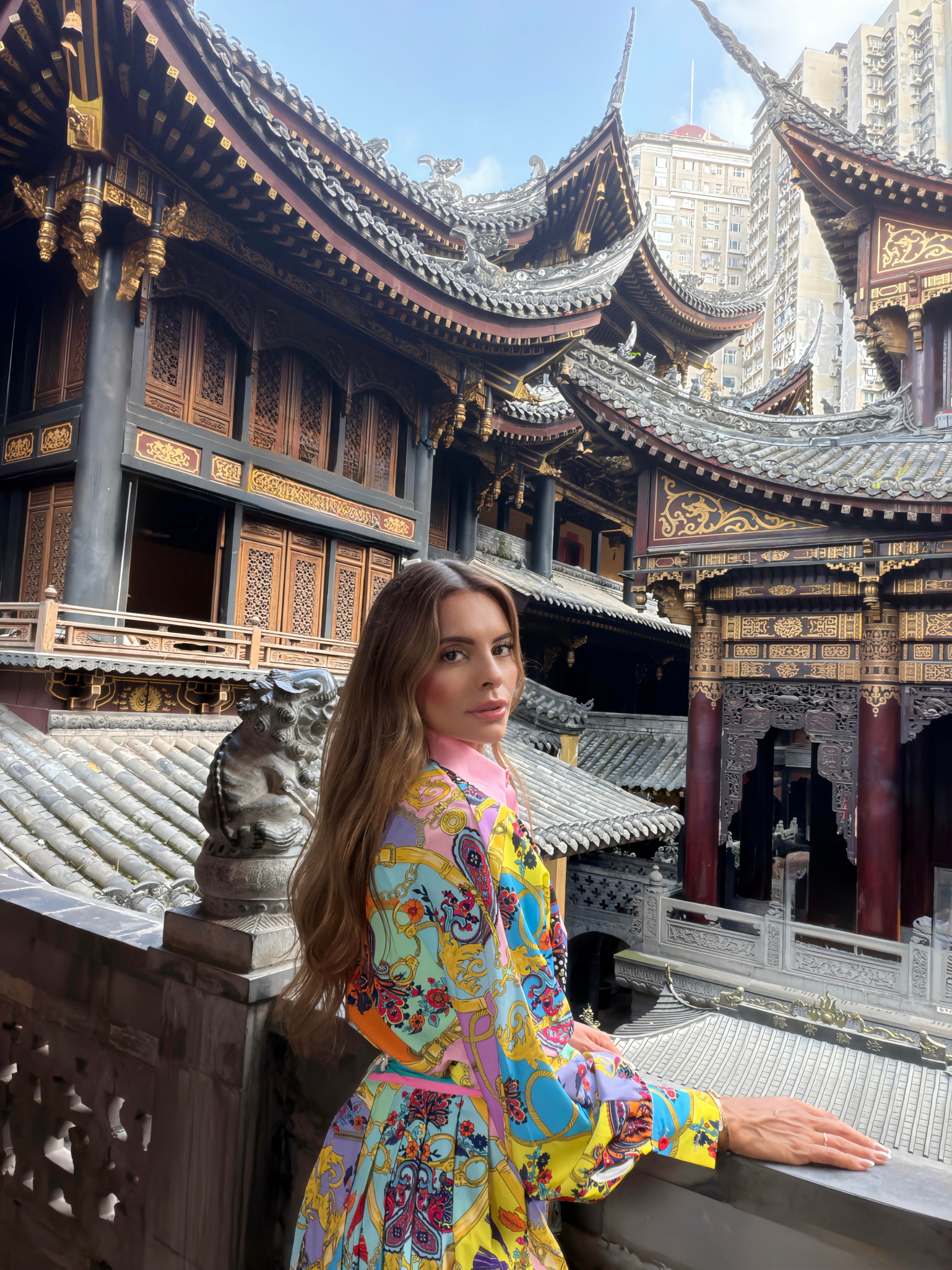 Young woman with long hair in colorful traditional attire stands in front of ornate Asian temple architecture.