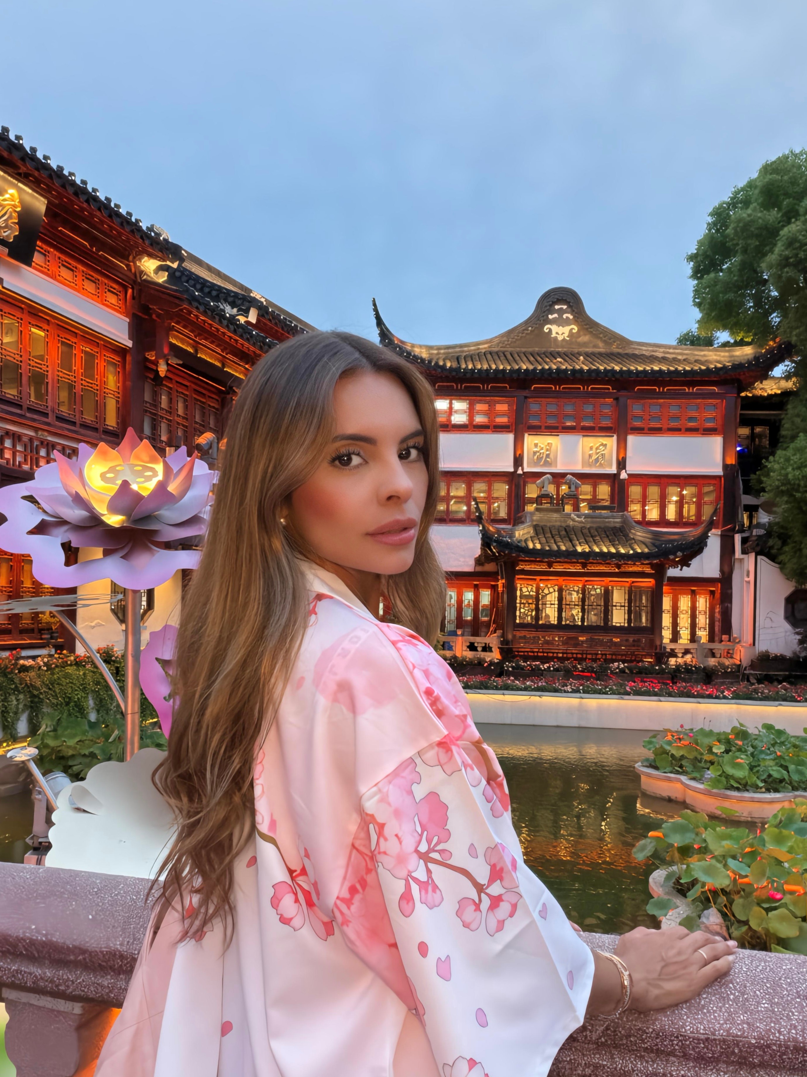 Young woman with long hair in a pink and white kimono standing near a pond with lotus flowers, traditional Asian architecture in background.