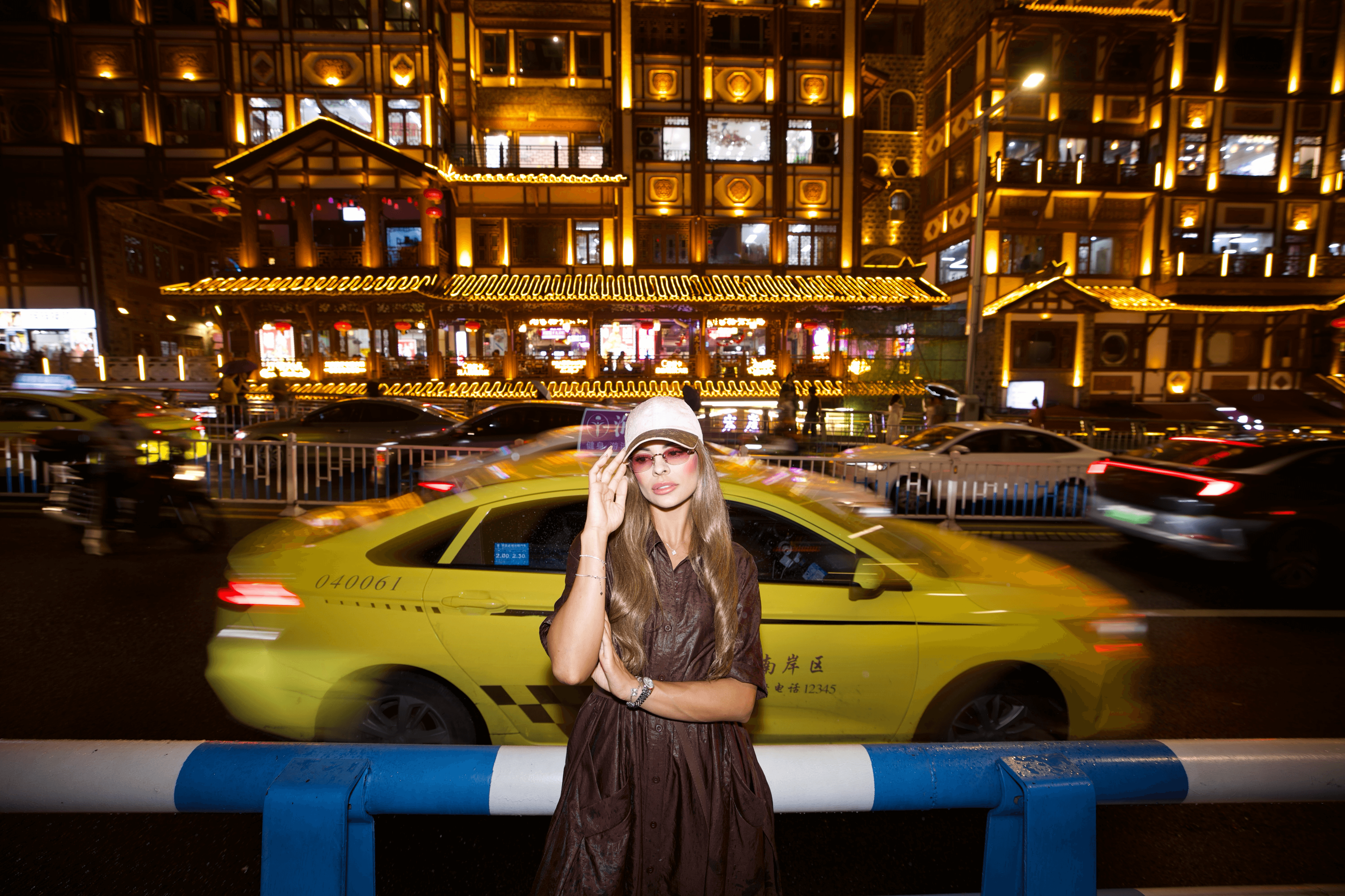 Young woman with long hair and glasses standing in front of a city street at night, yellow taxi and illuminated buildings behind her.