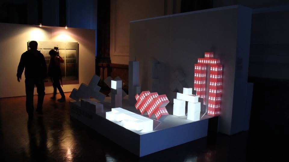 Exhibition with illuminated architectural models and people viewing artwork in a dimly lit gallery.