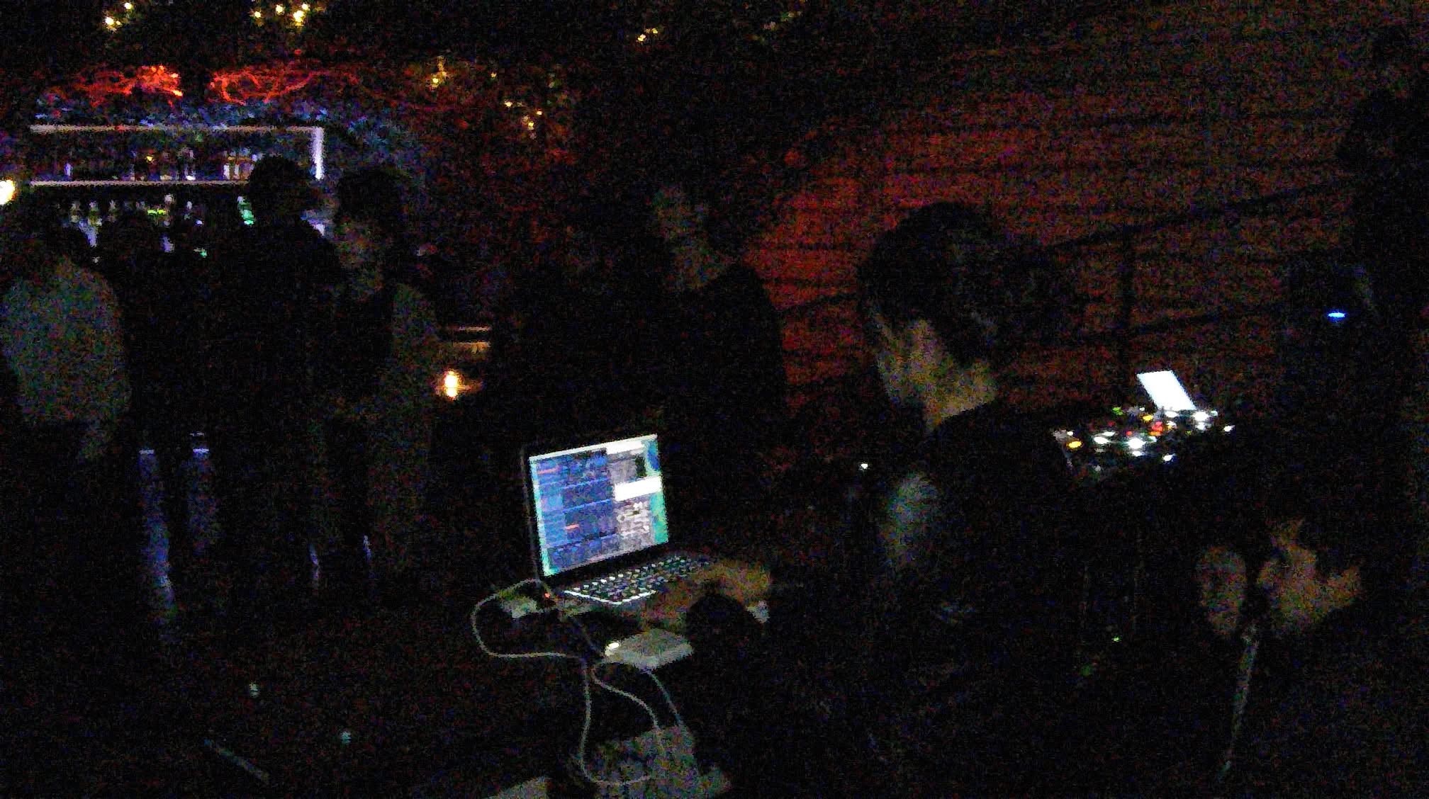 Person operating DJ equipment with a laptop in a dark, crowded venue with people in the background.