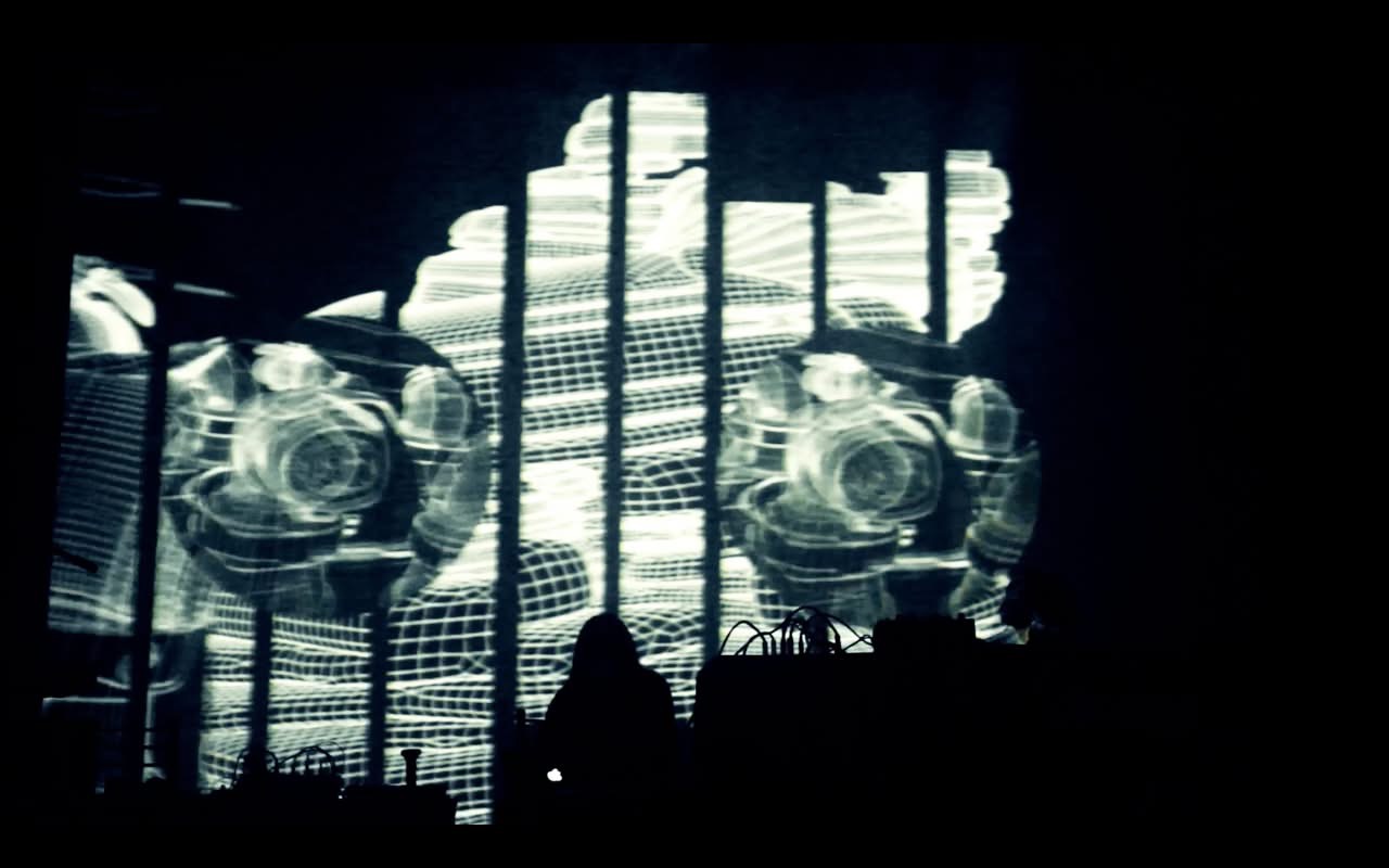 Silhouettes of people in front of a large screen displaying abstract visuals, with bright light and dark shadows.