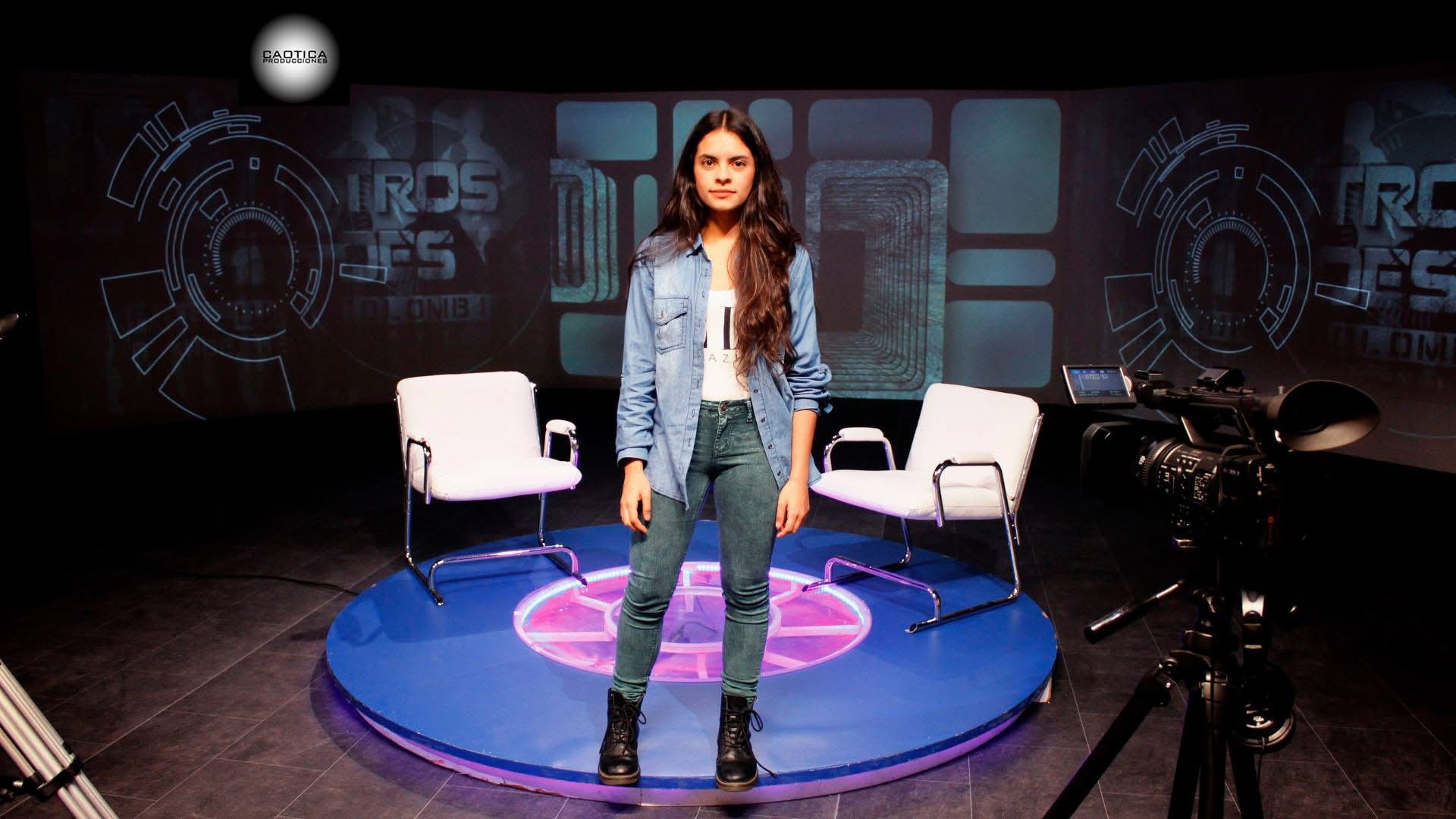 Woman standing on a circular stage with chairs, a camera, and a dark background with graphics.