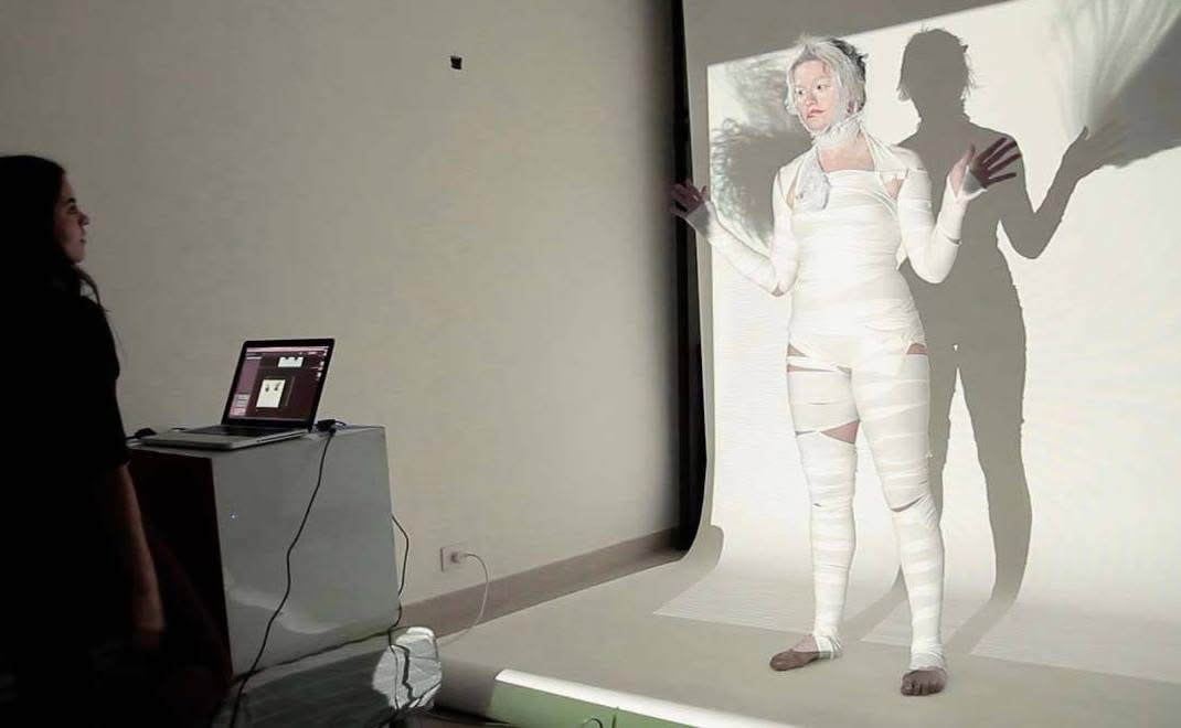 Person standing in front of a projection with a shadow cast on the wall, in a room with a laptop on a table.
