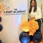 Woman standing next to a sign for The Light Academy, holding a cluster of yellow and black balloons, smiling.