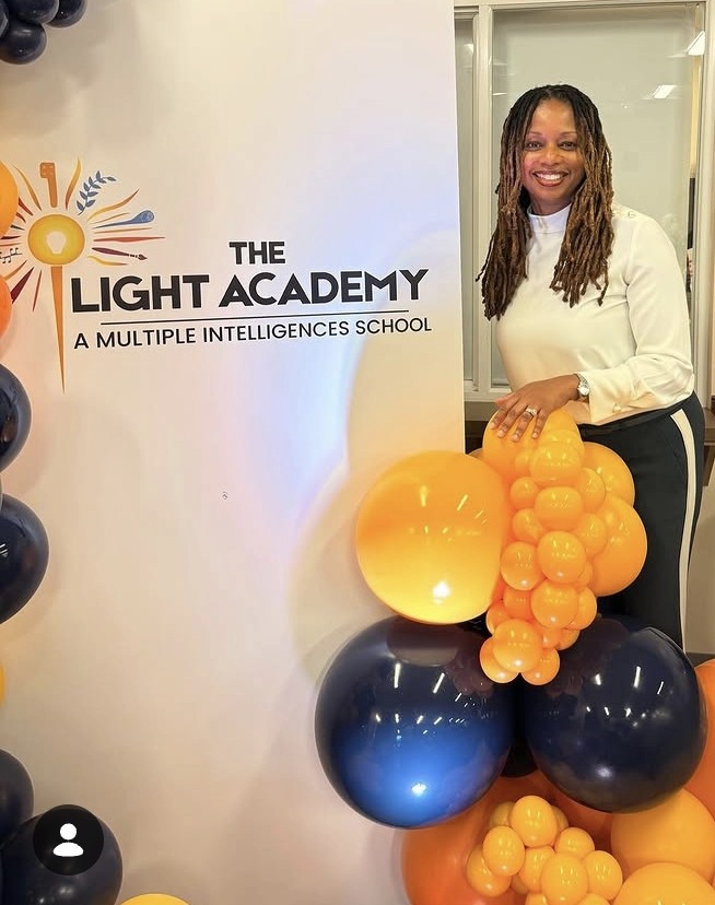 Woman standing next to a sign for The Light Academy, holding a cluster of yellow and black balloons, smiling.