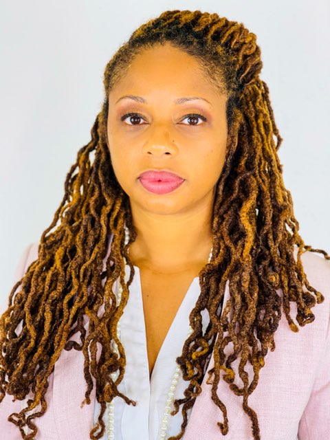 Portrait of a woman with long, styled dreadlocks, wearing a light-colored blazer, looking directly at the camera.
