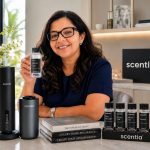 Woman with glasses smiling at a counter with scent products, books, and a black scentia sign in background.