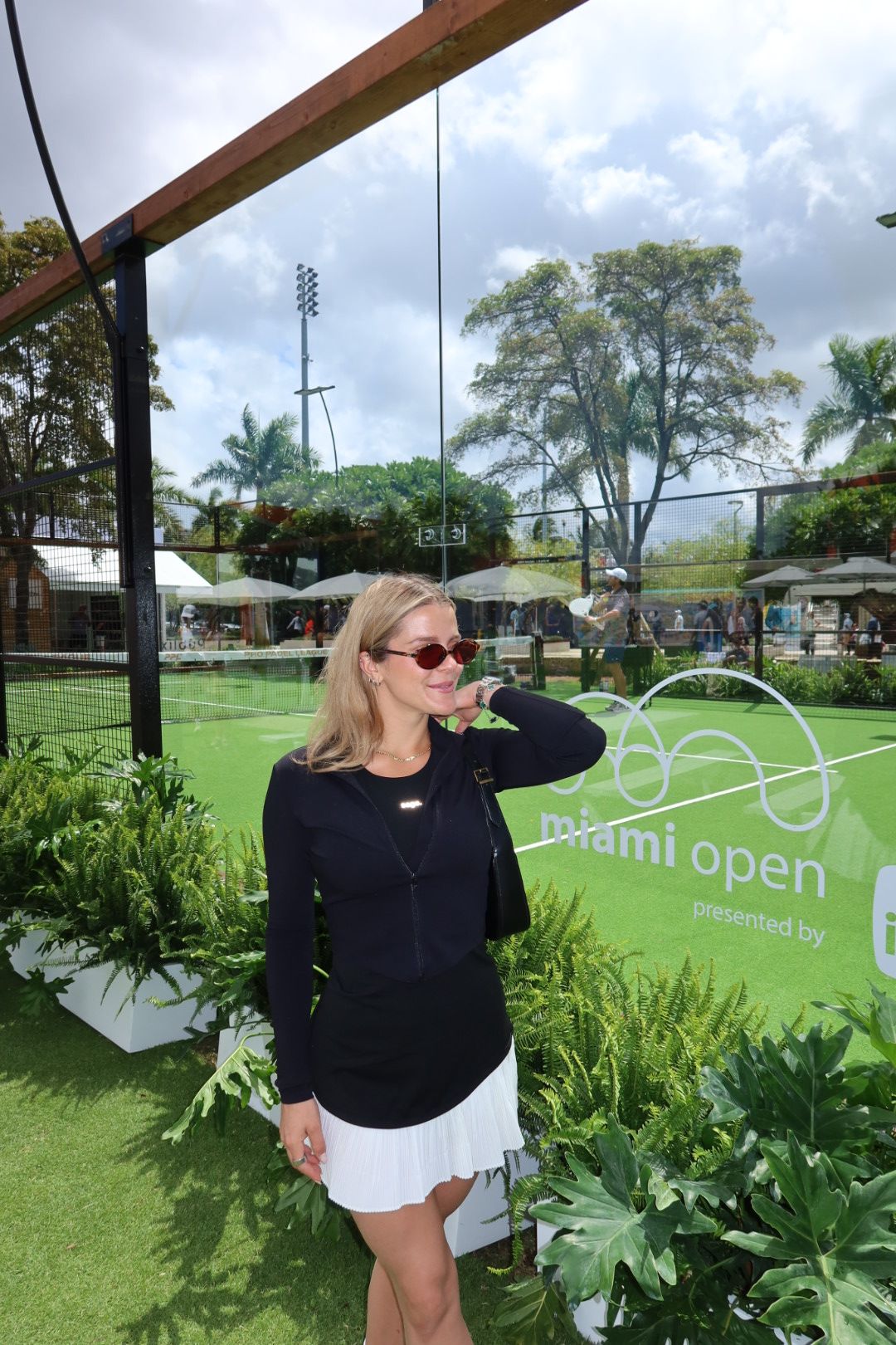 Woman with sunglasses and blonde hair standing on a green sports field, surrounded by plants and trees, with a cloudy sky.