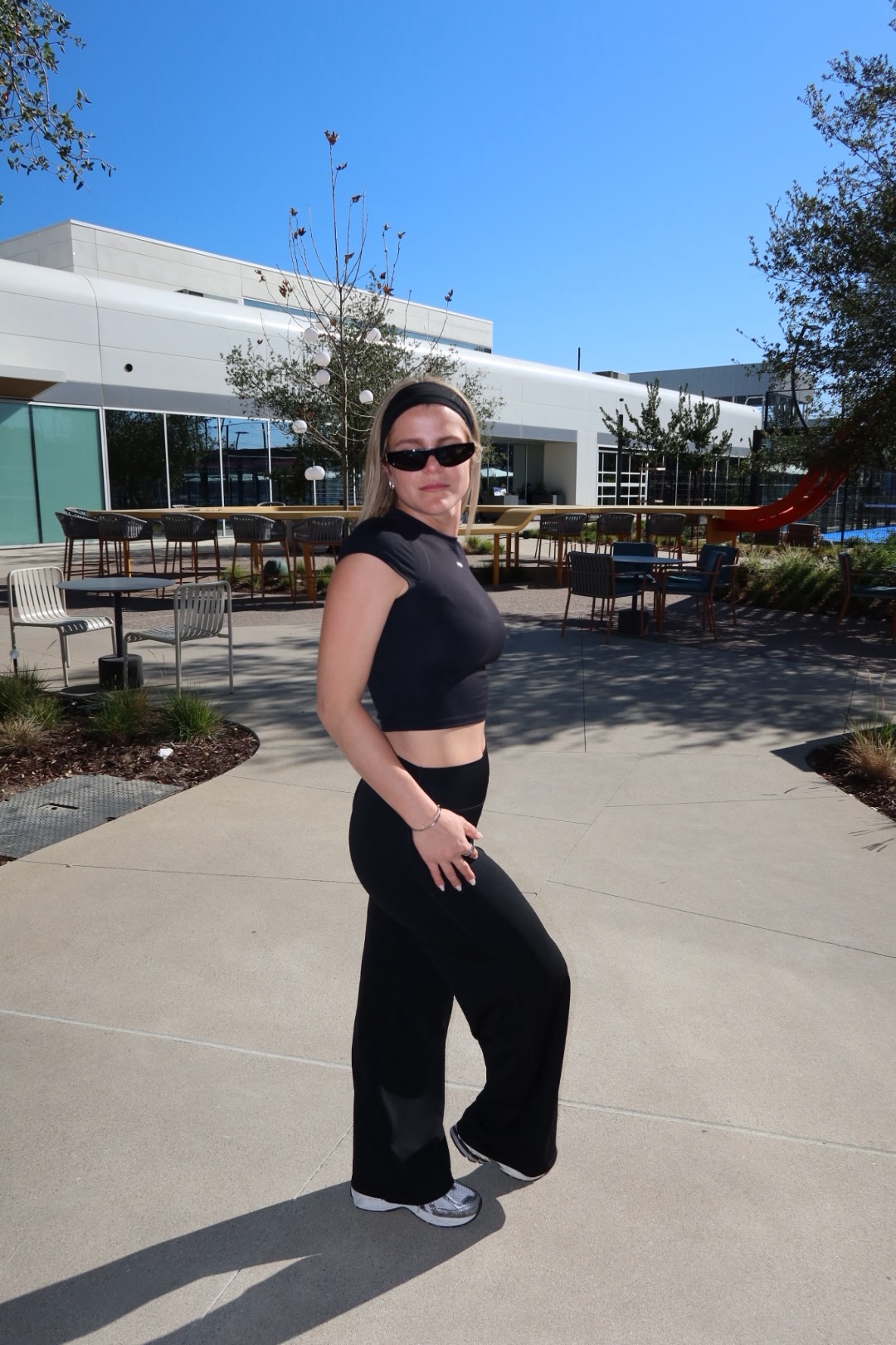 Woman in black outfit standing outdoors on a sunny day with modern building and trees in background.