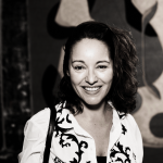 Smiling woman with curly hair wearing a patterned blouse, standing in front of abstract artwork.