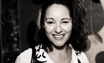 Smiling woman with curly hair wearing a patterned blouse, standing in front of abstract artwork.