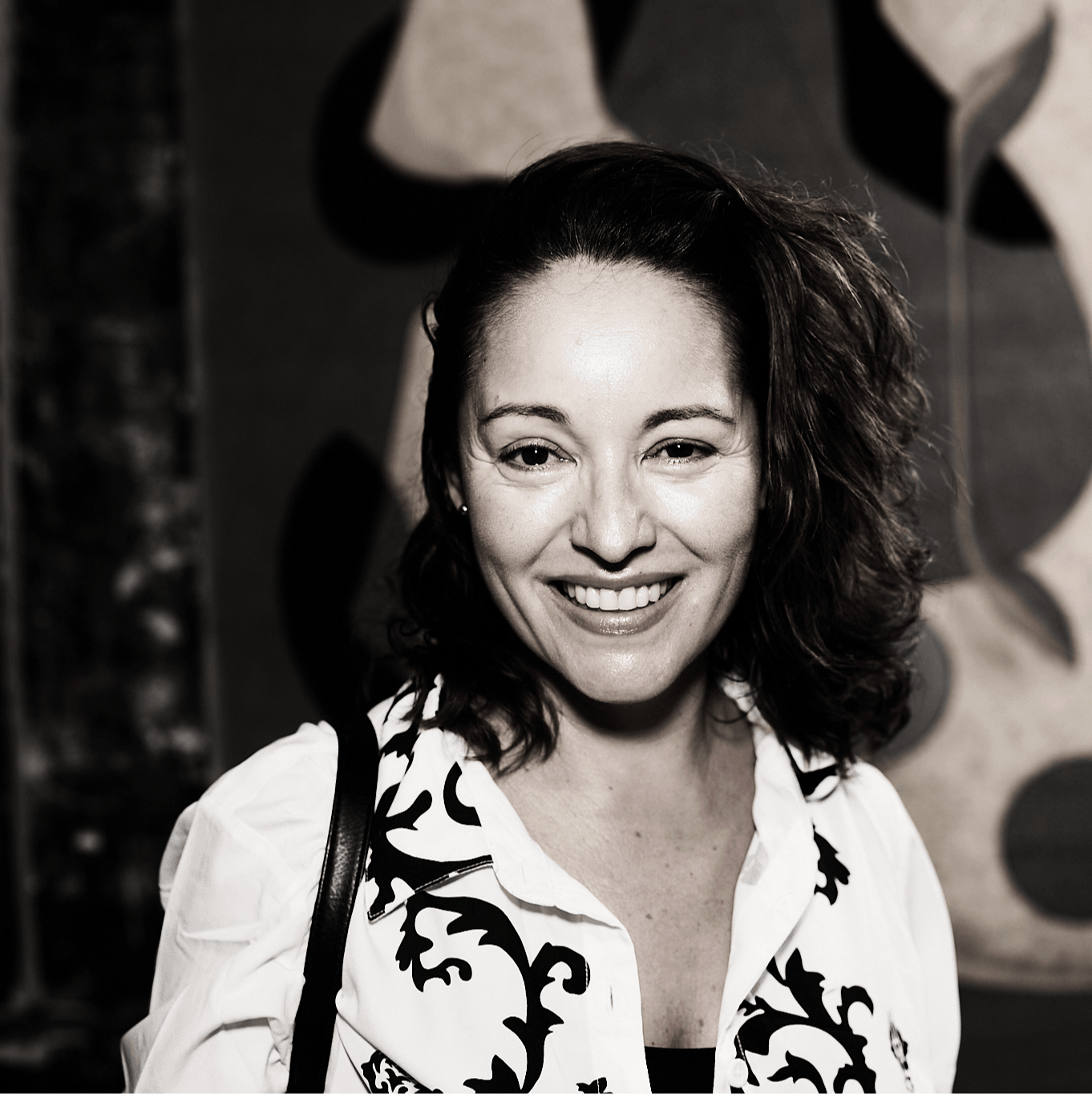 Smiling woman with curly hair wearing a patterned blouse, standing in front of abstract artwork.