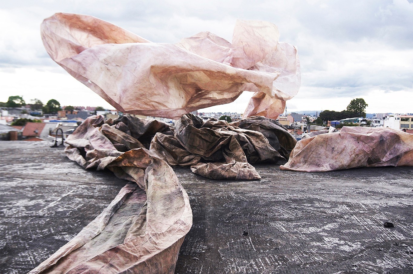 Large abstract sculpture made of pink and brown materials on a rooftop with a cityscape background.