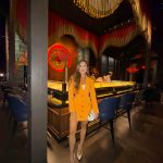 Woman in yellow blazer dress standing in a dimly lit restaurant with colorful decor and seating.