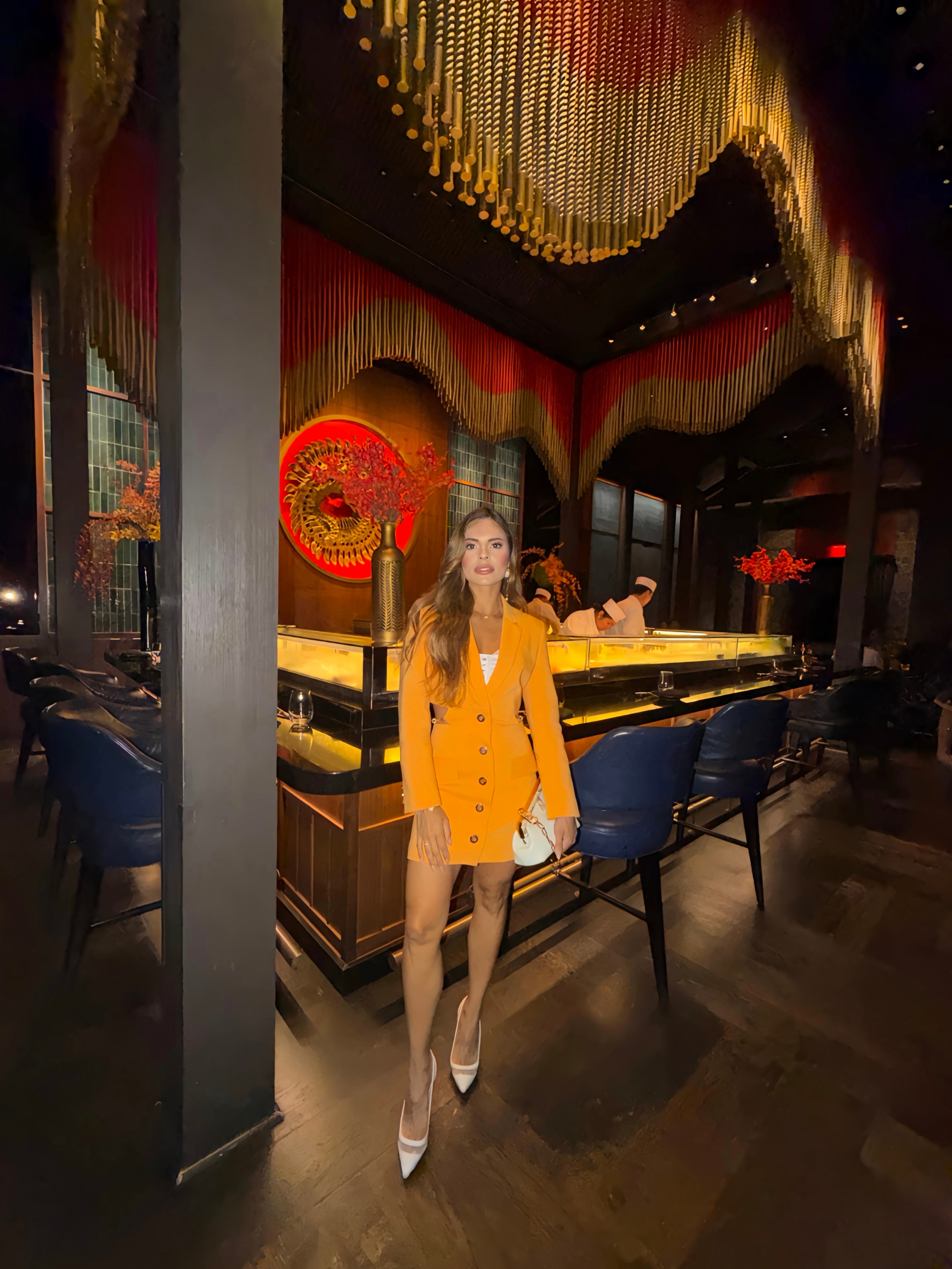 Woman in yellow blazer dress standing in a dimly lit restaurant with colorful decor and seating.