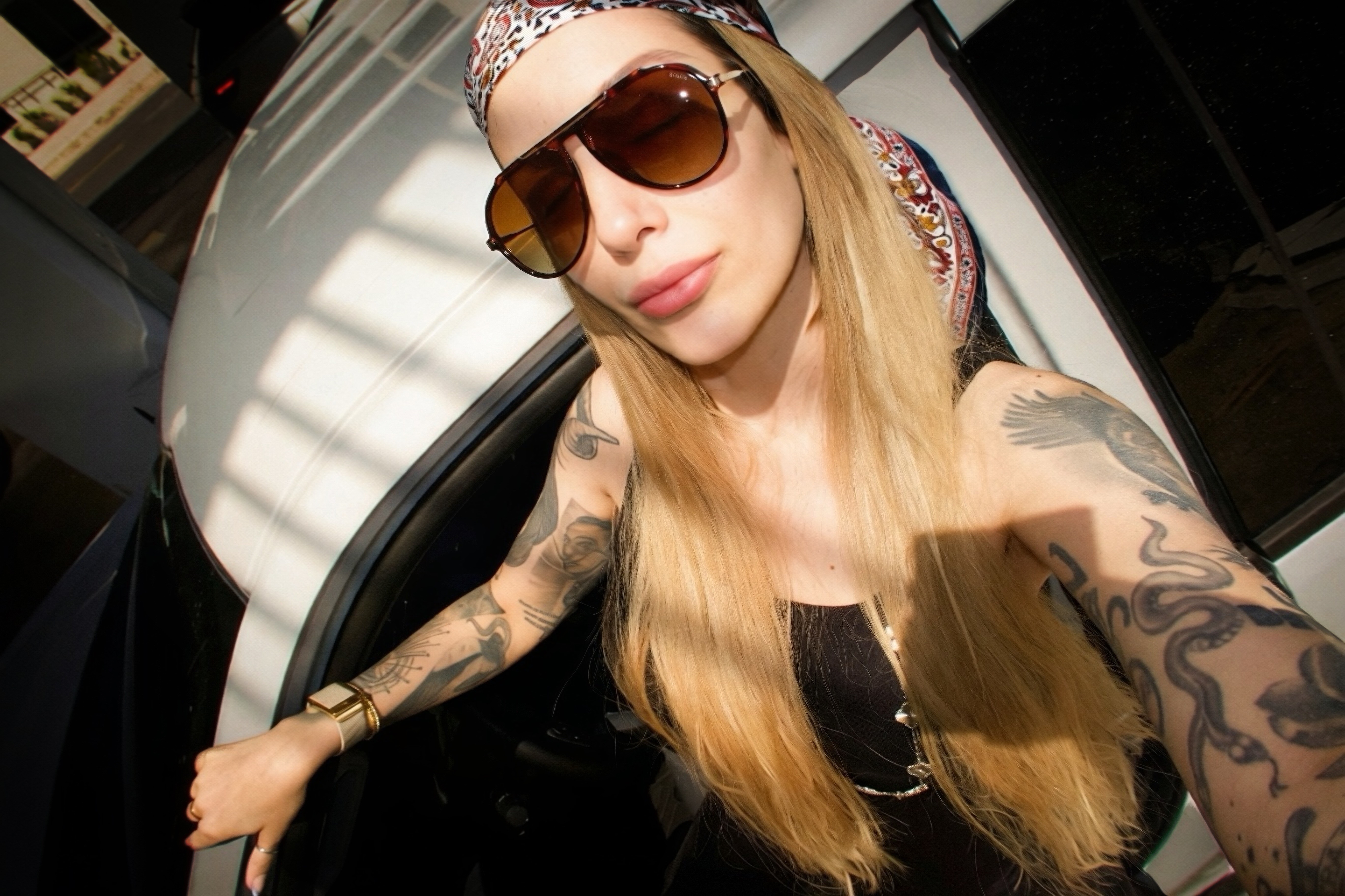 Woman with long blonde hair wearing sunglasses, a headscarf, and tattoos, taking a selfie indoors.