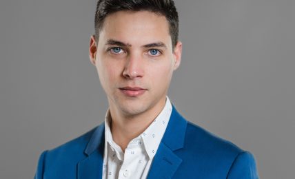 Young man with dark hair and blue eyes wearing a blue blazer and white shirt against a gray background.