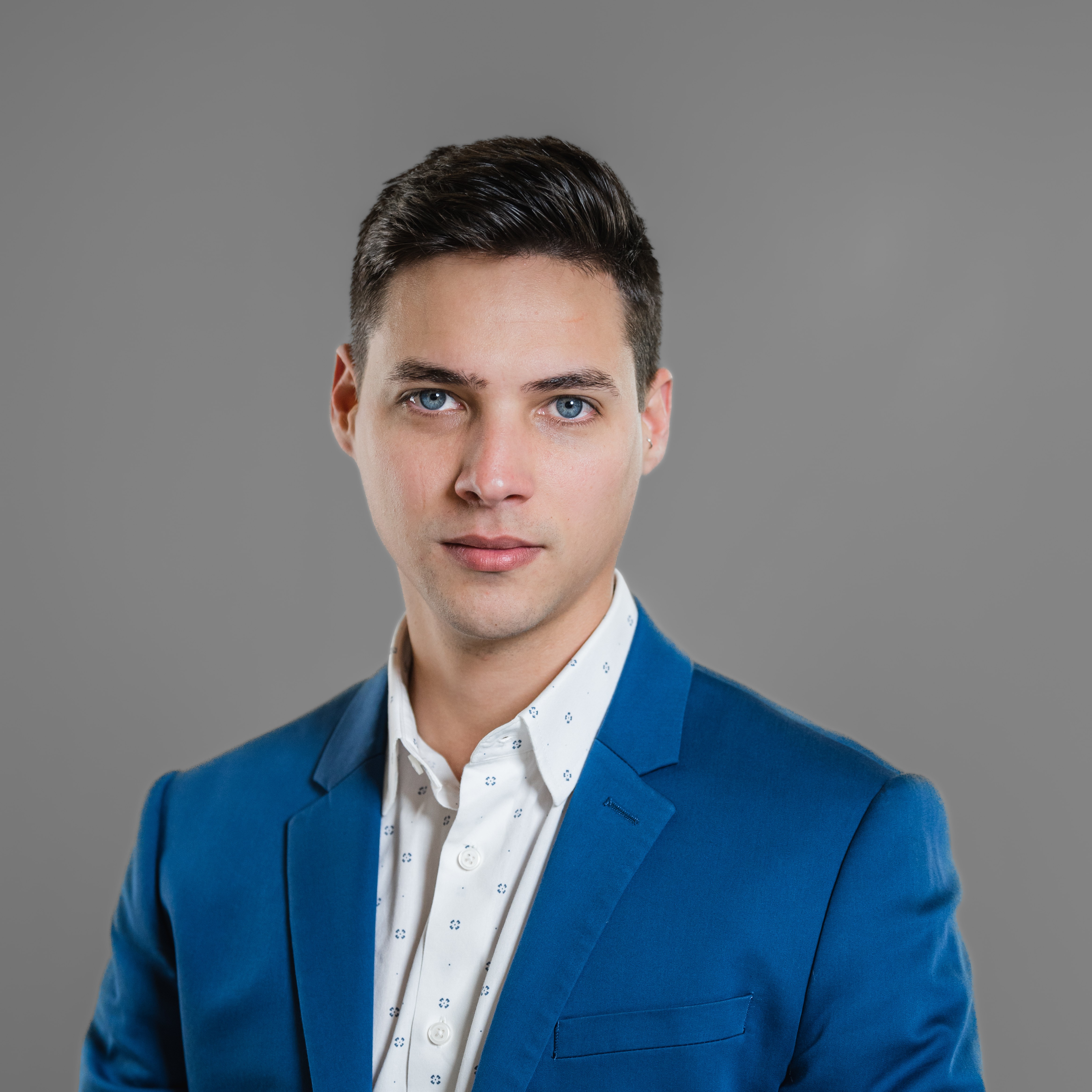 Young man with dark hair and blue eyes wearing a blue blazer and white shirt against a gray background.
