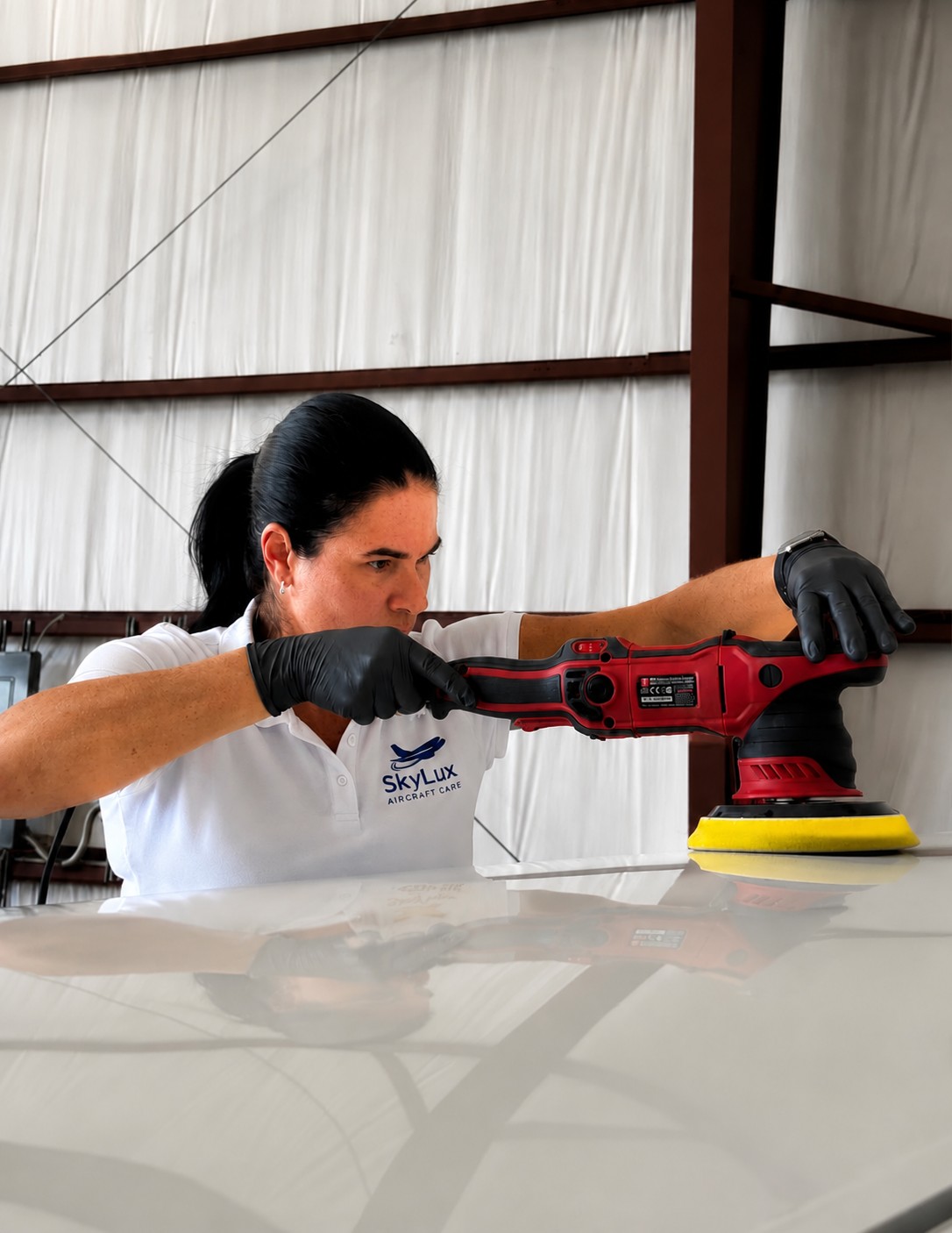 Woman using a red polishing machine on a white surface, wearing black gloves and a white shirt, in a warehouse setting.