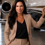Woman in beige suit standing next to private jet in hangar, looking at camera.