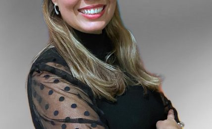 Woman with long hair smiling, wearing a black top with sheer polka-dot sleeves, standing against a plain background.