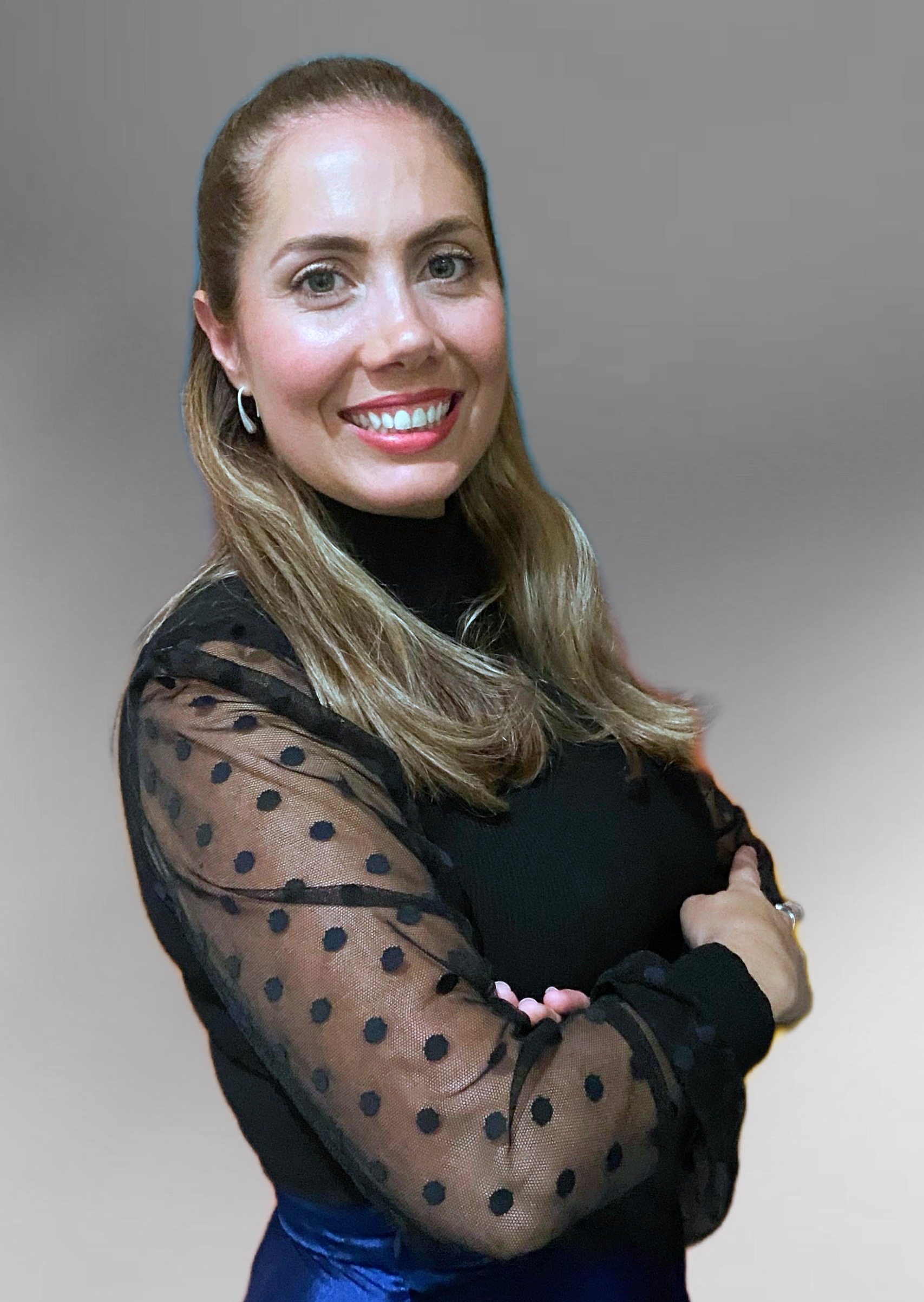 Woman with long hair smiling, wearing a black top with sheer polka-dot sleeves, standing against a plain background.