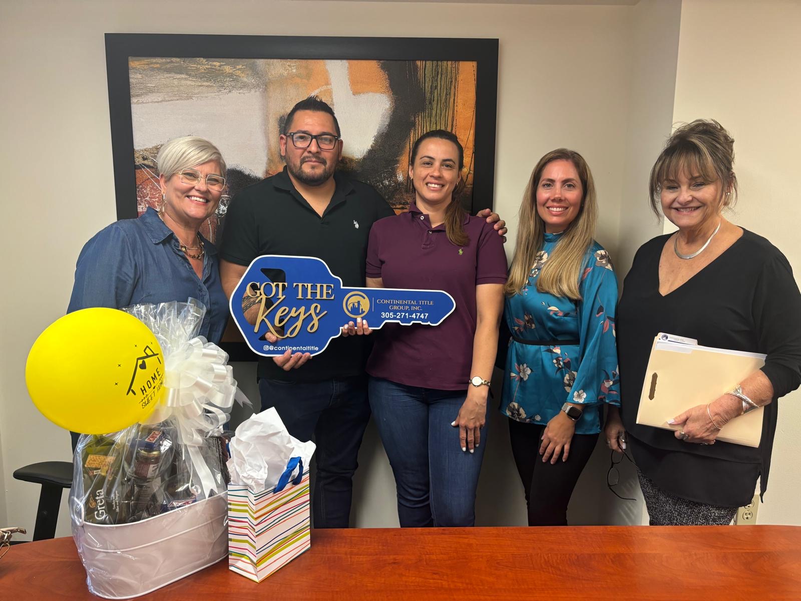 Group of five people standing indoors, holding a blue sign, smiling, with gift baskets and a yellow balloon on a table.