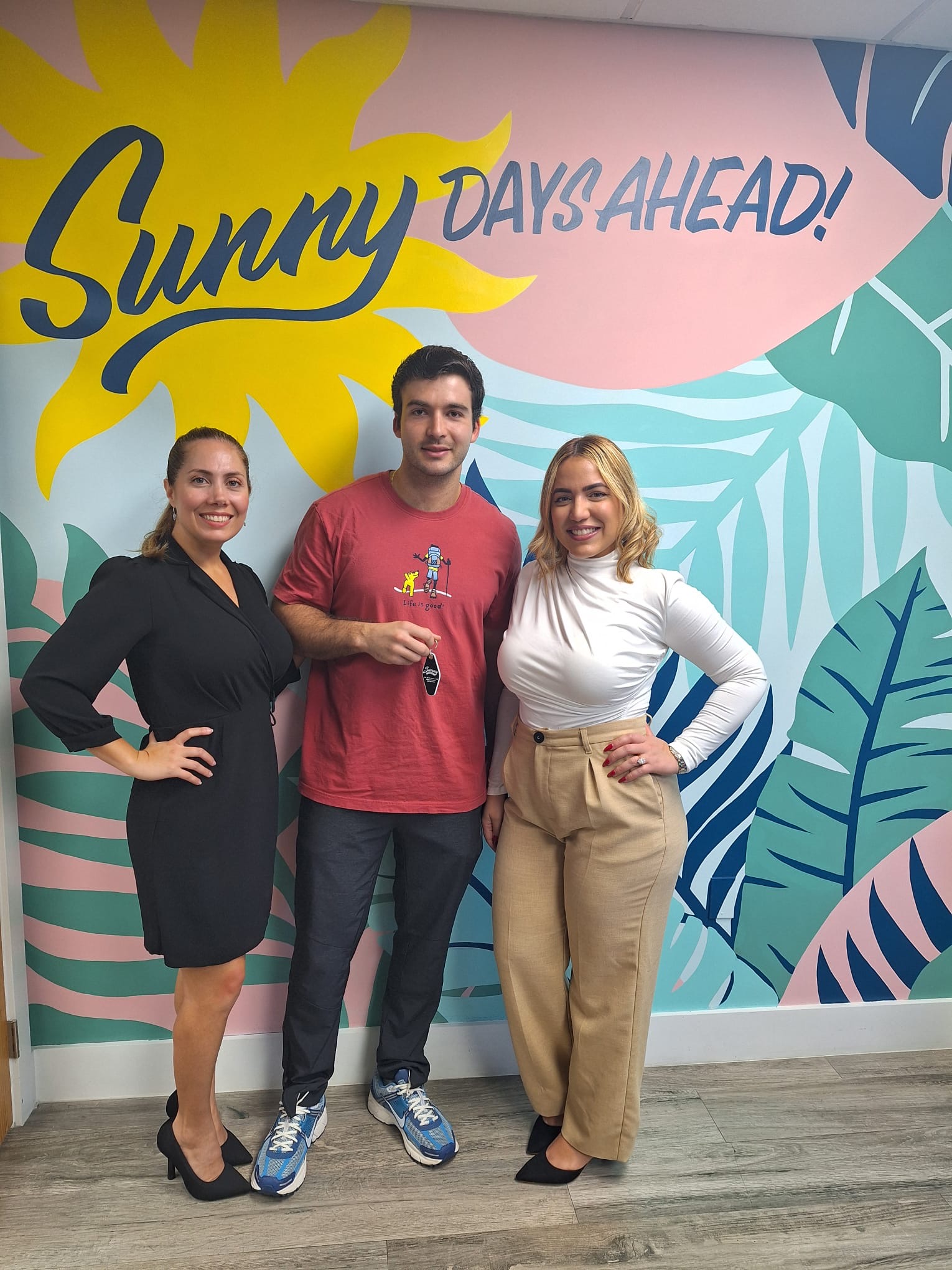 Three people standing in front of a colorful mural with a sun and leaves, smiling, with the text 'Sunny Days Ahead!'.