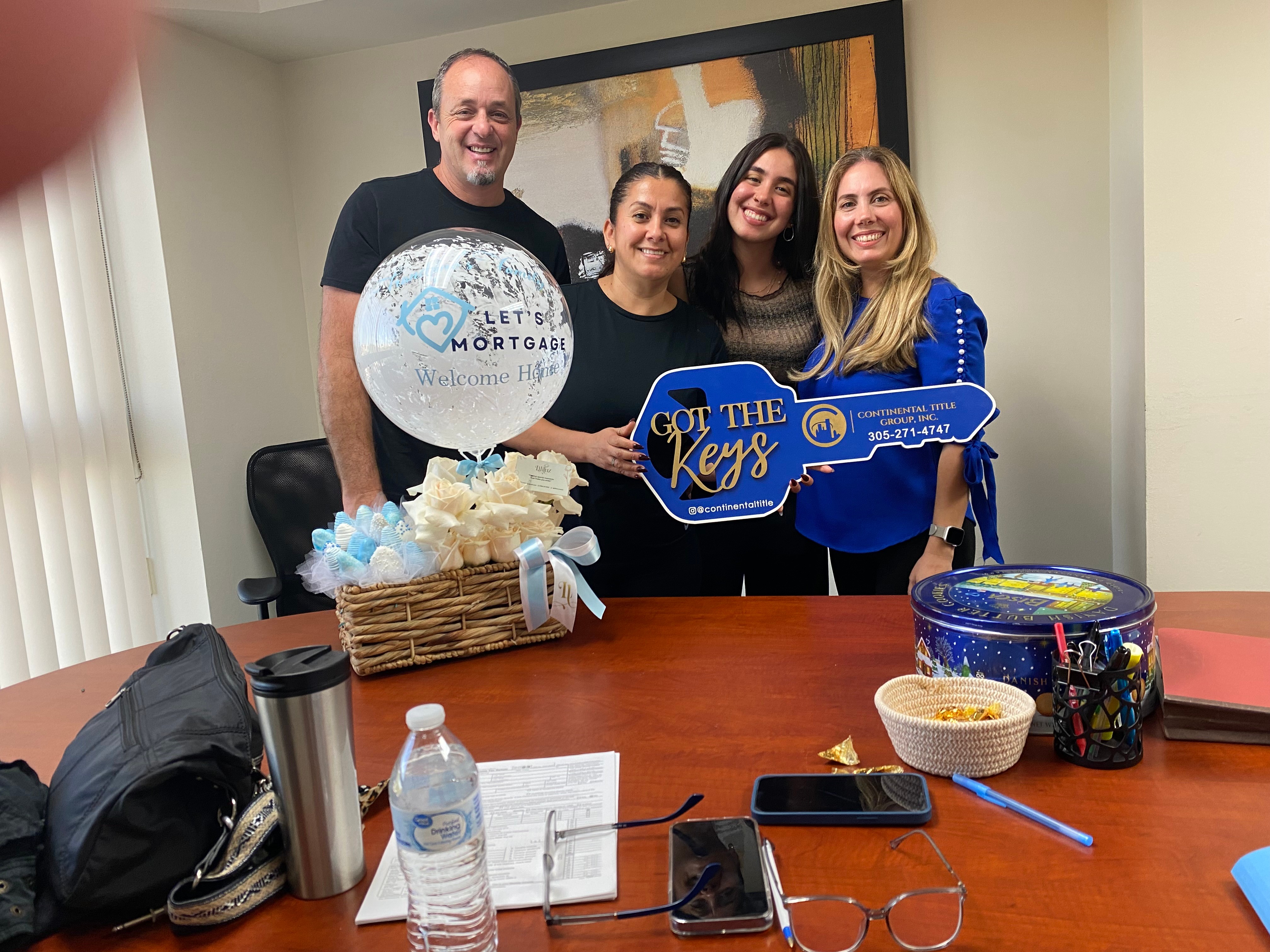 Four people smiling, holding a large key and a balloon, standing behind a table with gifts and snacks, in a room with a large window.