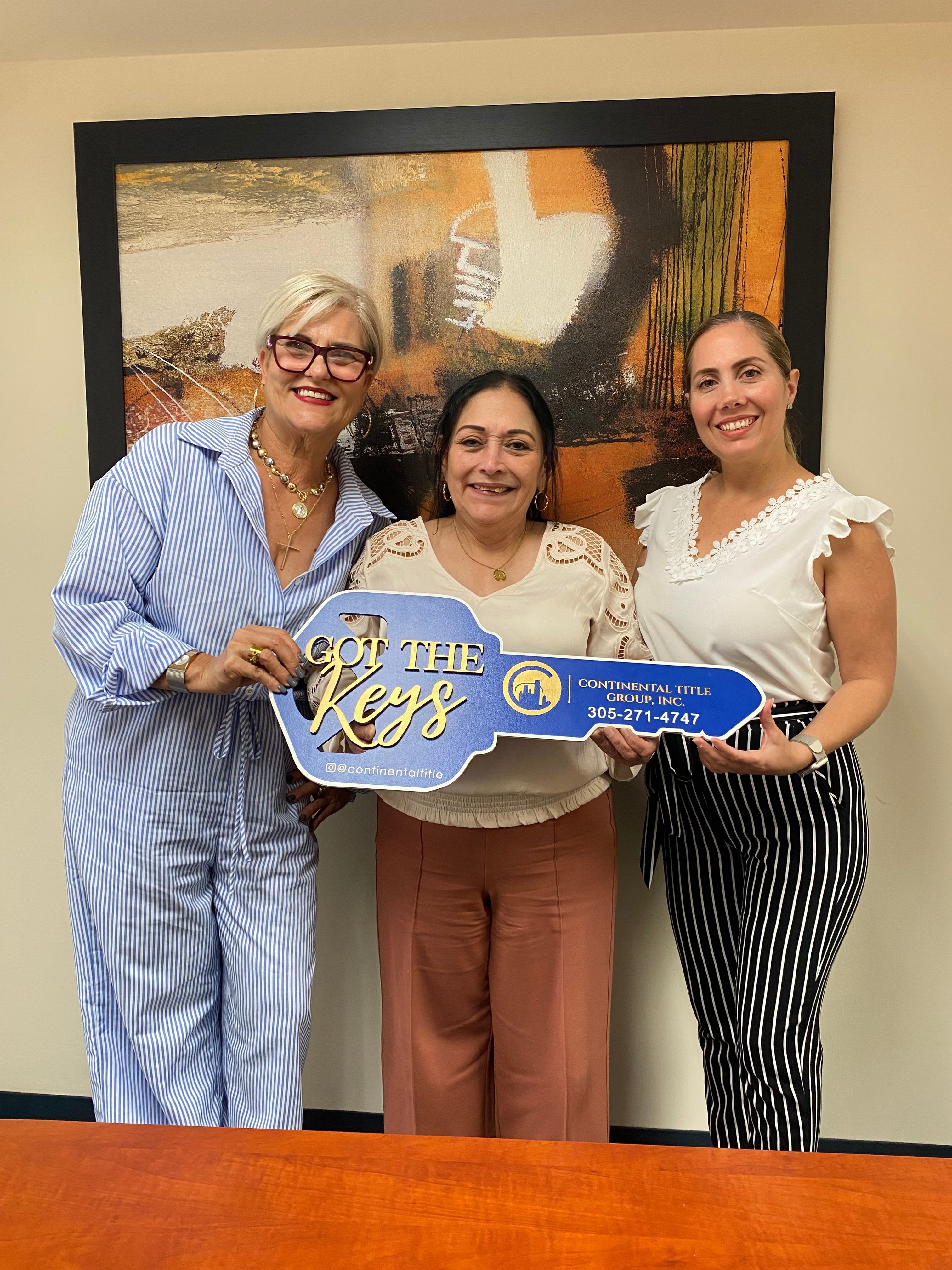 Three women standing indoors, holding a sign that says 'The Big Day,' smiling at the camera.