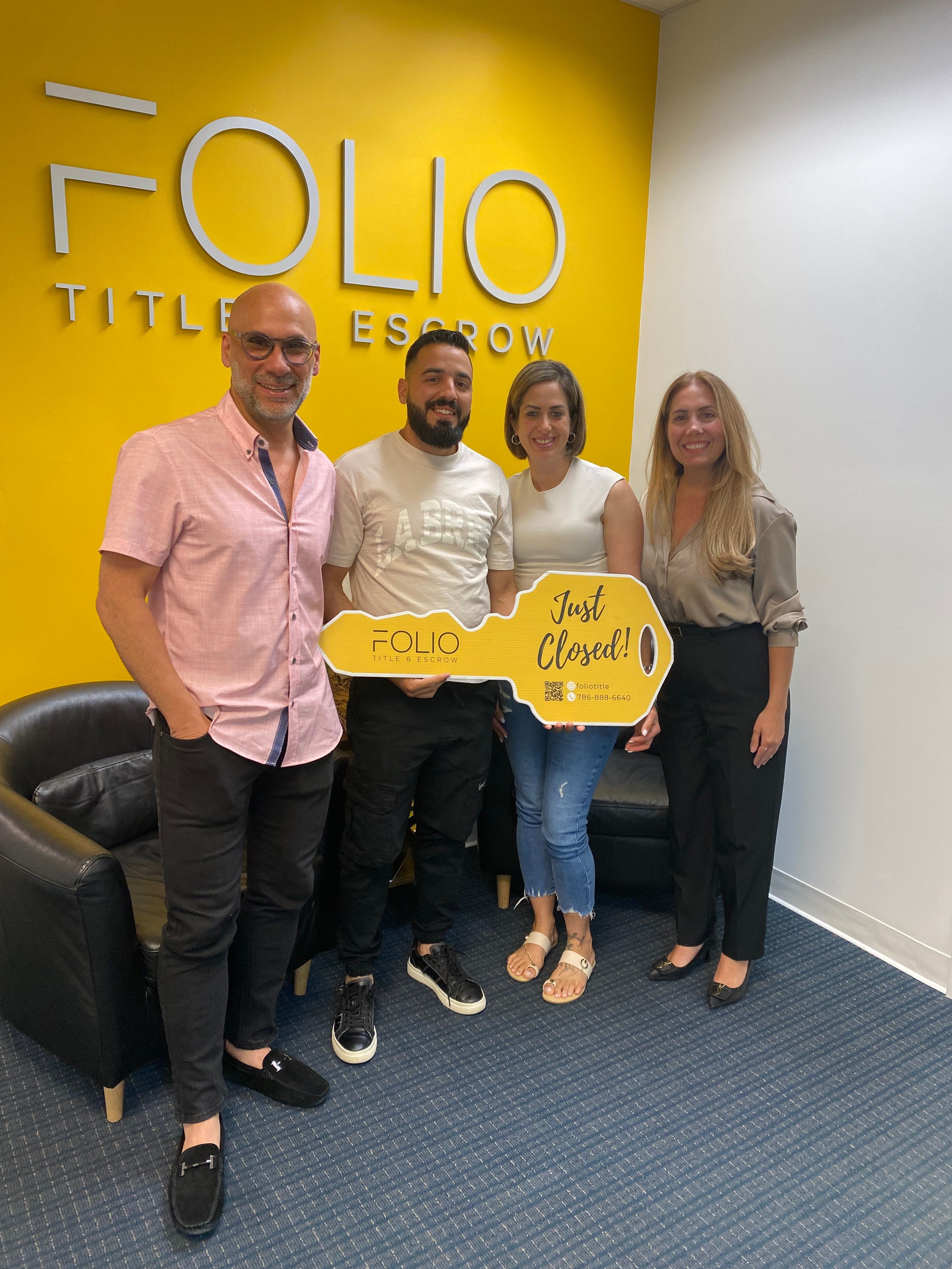 Four people standing in a hallway with a yellow wall and blue carpet, holding a yellow sign with black text.