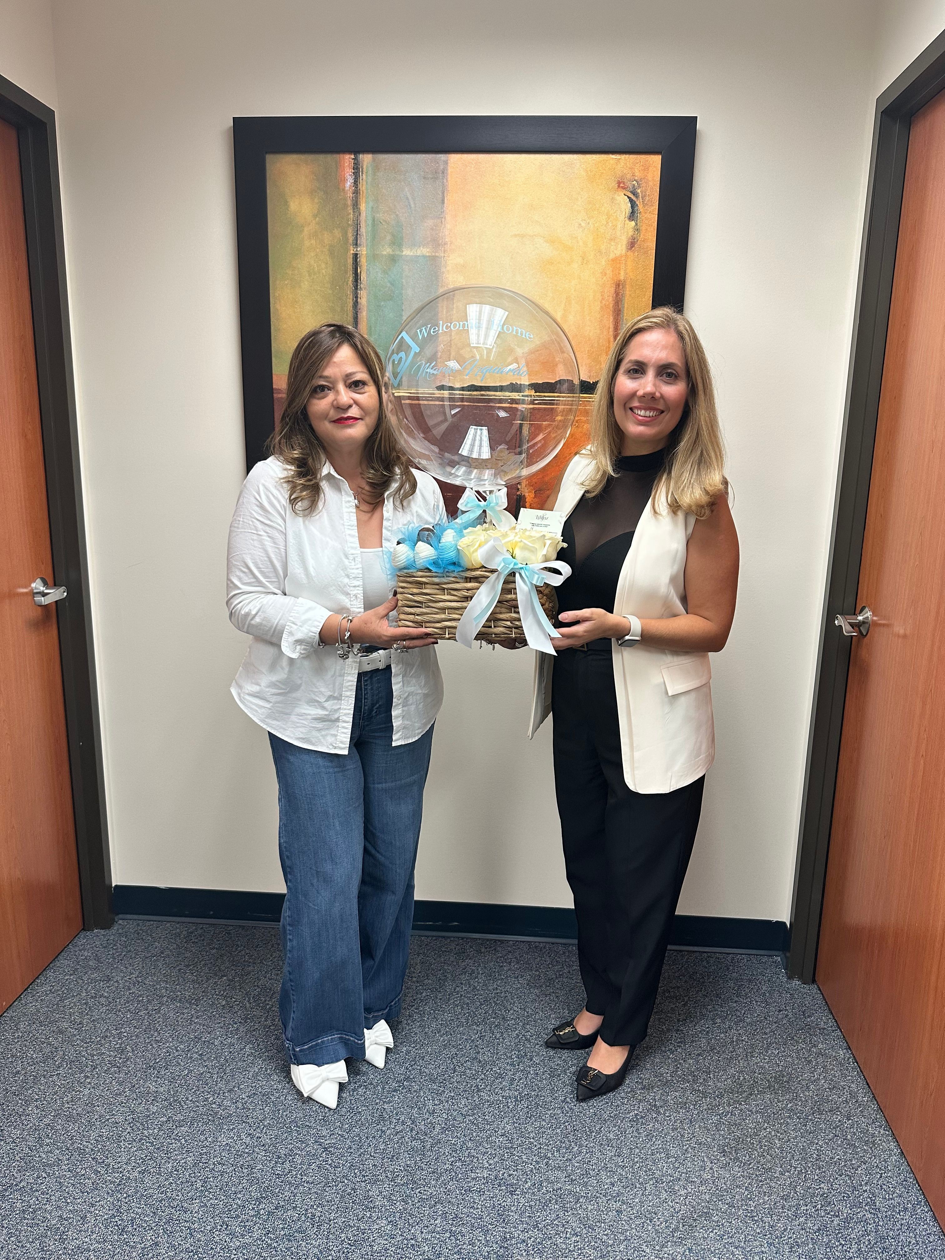 Two women standing in an office hallway, one holding a gift basket with a clear balloon, in front of a framed painting.