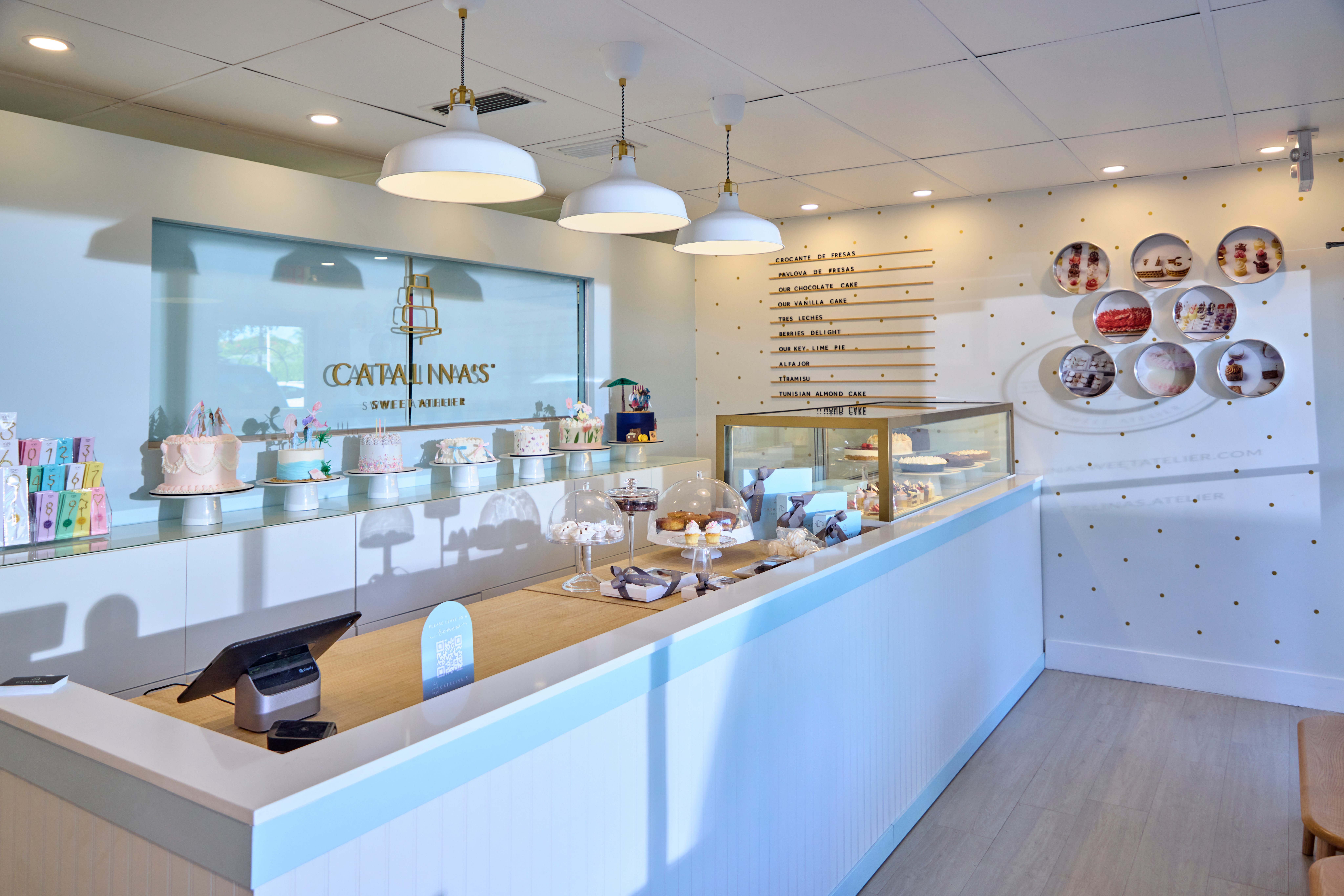 Interior of a modern bakery or cafe with a white counter, display case, and decorative plates on the wall, illuminated by hanging lights.